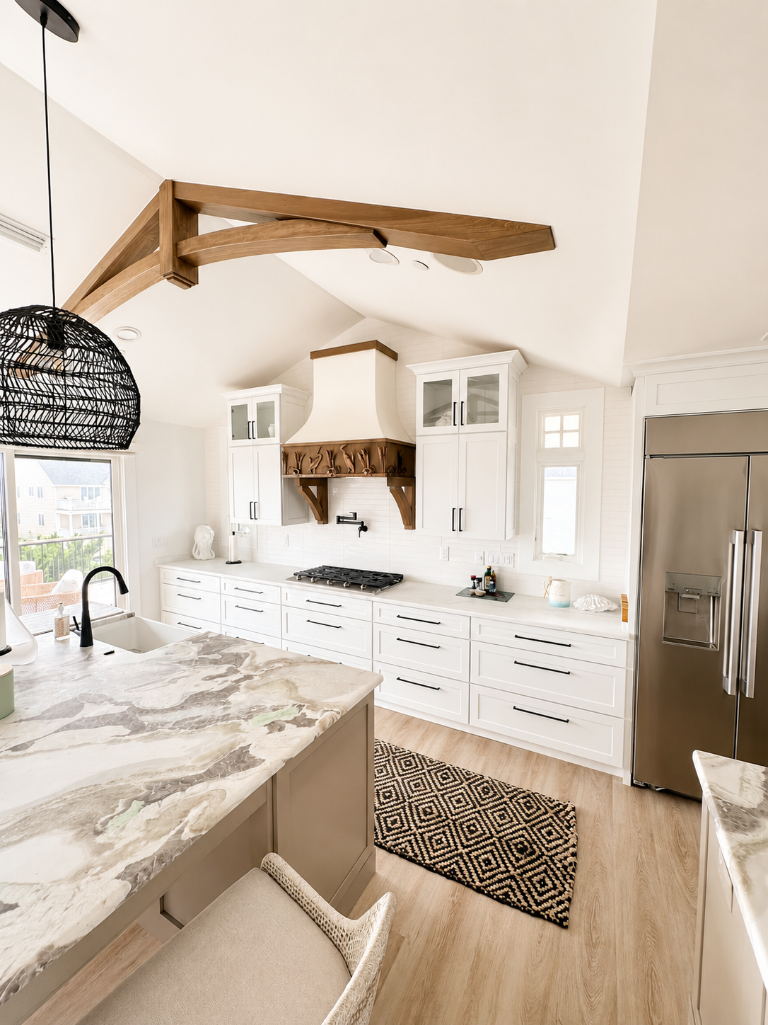 Modern kitchen with white cabinets, marble island, stainless steel refrigerator, wooden beams on ceiling, and a black patterned rug on light wood flooring.