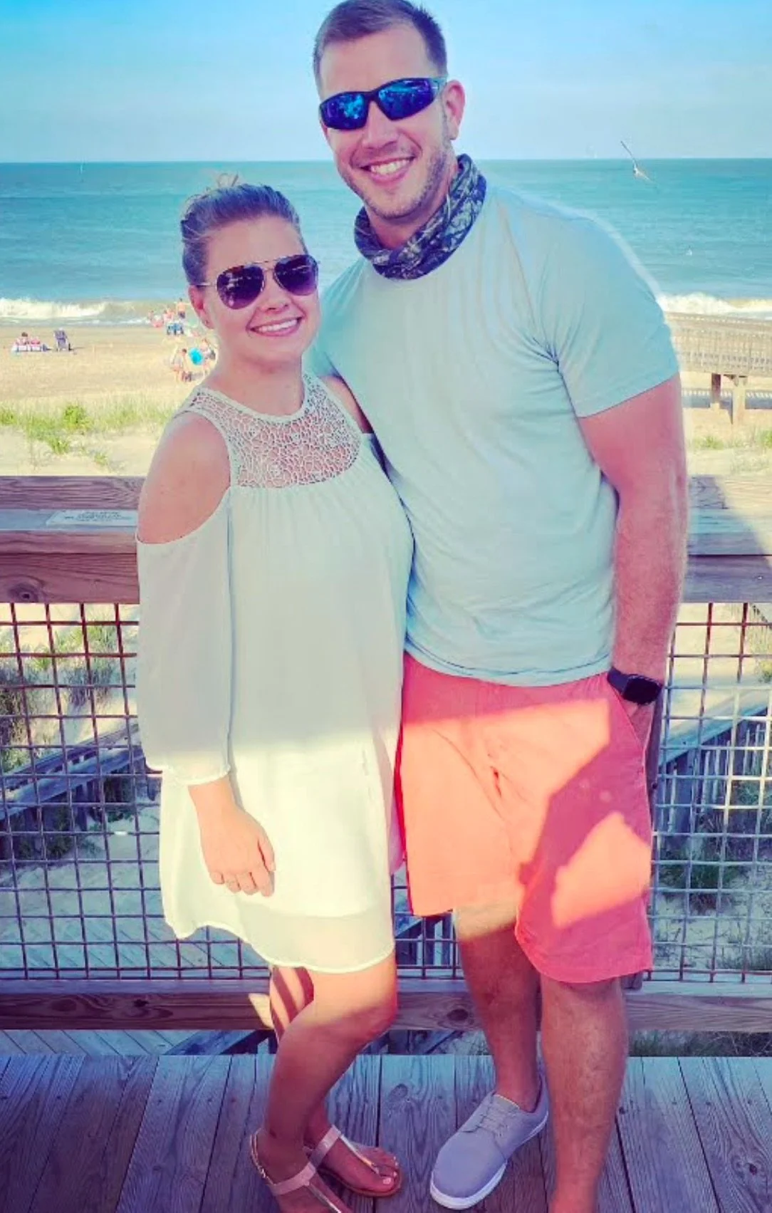 A man and woman smiling together on a beach boardwalk. The woman wears sunglasses and a white dress with open shoulders, and the man wears sunglasses, a t-shirt, and coral shorts.