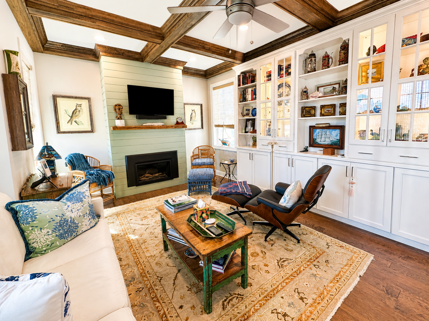 Living room with wooden beams on ceiling, white wood-paneled walls, a beige couch with floral pillows, a wooden coffee table, a black leather lounge chair with a wooden frame, a blue armchair, a fireplace with a flat-screen TV above, white shelving with decorative items, a large patterned rug, and large windows with blinds.