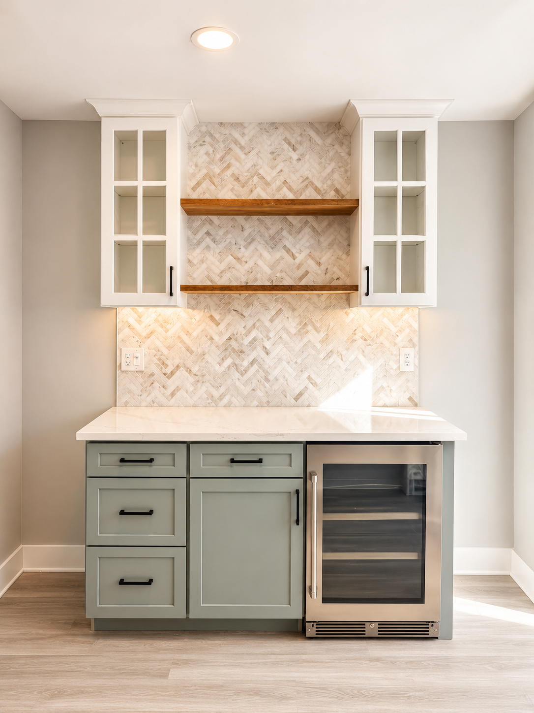 Kitchen area with light gray cabinets, a white countertop, and two upper glass-front cabinets. There are two wooden shelves against a herringbone-patterned tile backsplash, with outlets on either side of the counter. A small wine fridge is built into the lower cabinetry.
