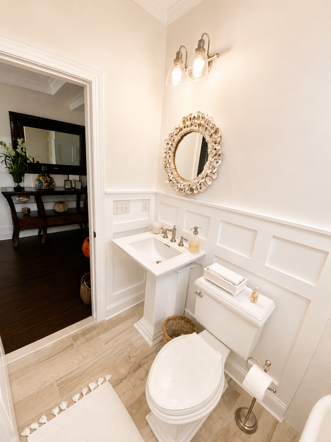 A small bathroom with white walls, a white toilet, and a white sink with chrome fixtures. Above the sink is a decorative oval mirror with a shell frame. A three-light wall sconce with exposed bulbs is mounted above the mirror. A wicker trash basket is beside the toilet. A white bathmat with tassels is on the beige wooden floor. In the background, through an open door, a dark-finished console table with various decorative items and a large mirror can be seen.