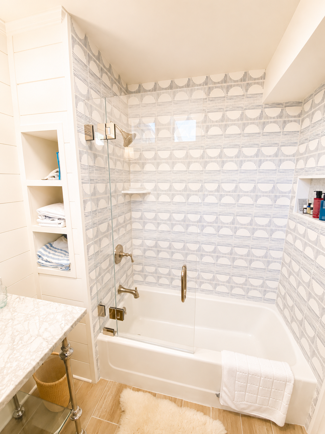 Bathroom with a bathtub, patterned tile walls, a glass shower door, built-in shelf with toiletries, an open cabinet with towels, a marble countertop, and a white rug.