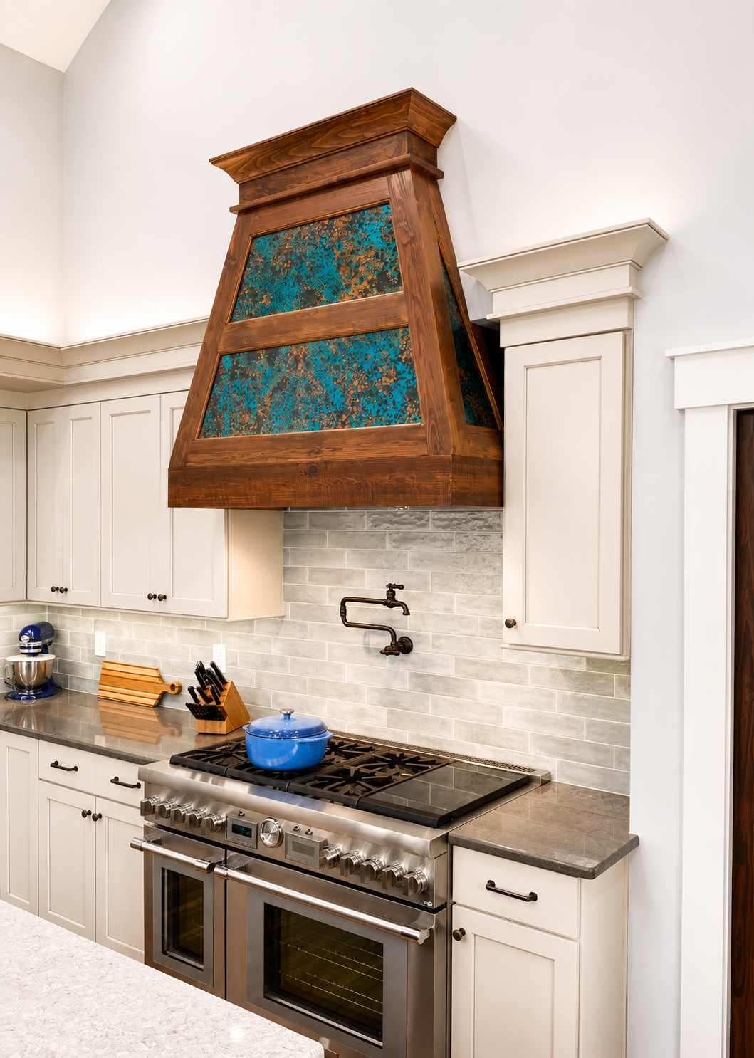 Kitchen with a stainless steel oven and stovetop, white cabinets, gray backsplash, and a decorative range hood with wood and blue patterned details.