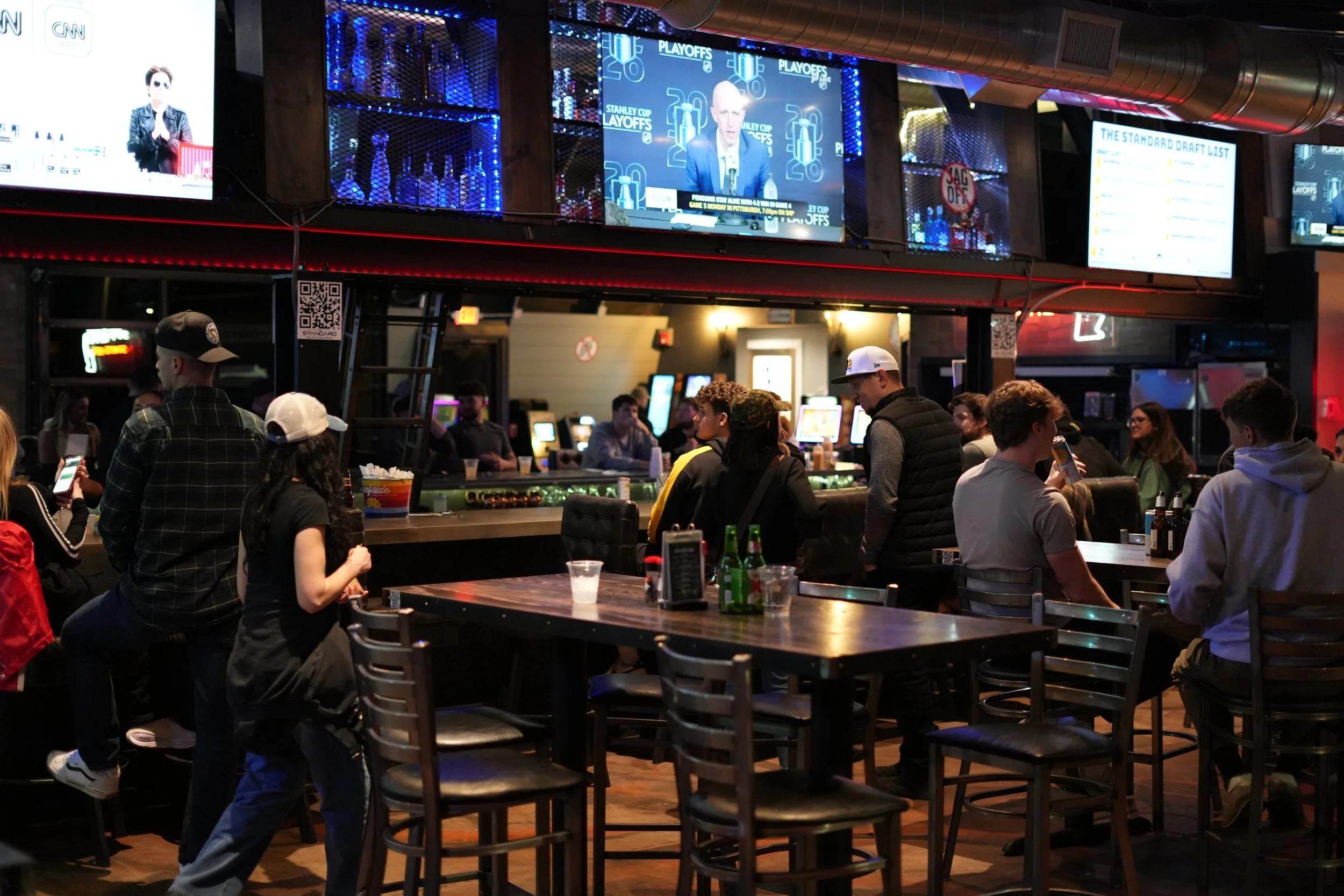 A busy bar scene with people gathered around tables and a bar counter, multiple large TV screens displaying sports and news, and neon lighting.