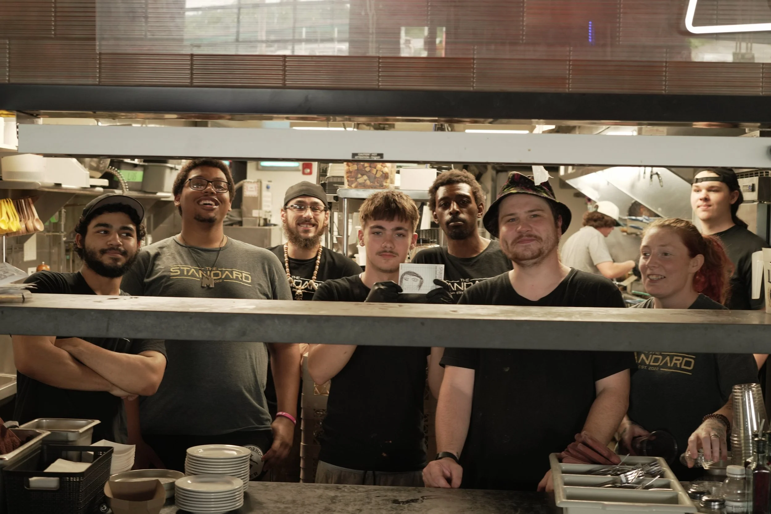 Group of people working behind a counter in a restaurant kitchen, some smiling, with plates and kitchen utensils in the foreground.
