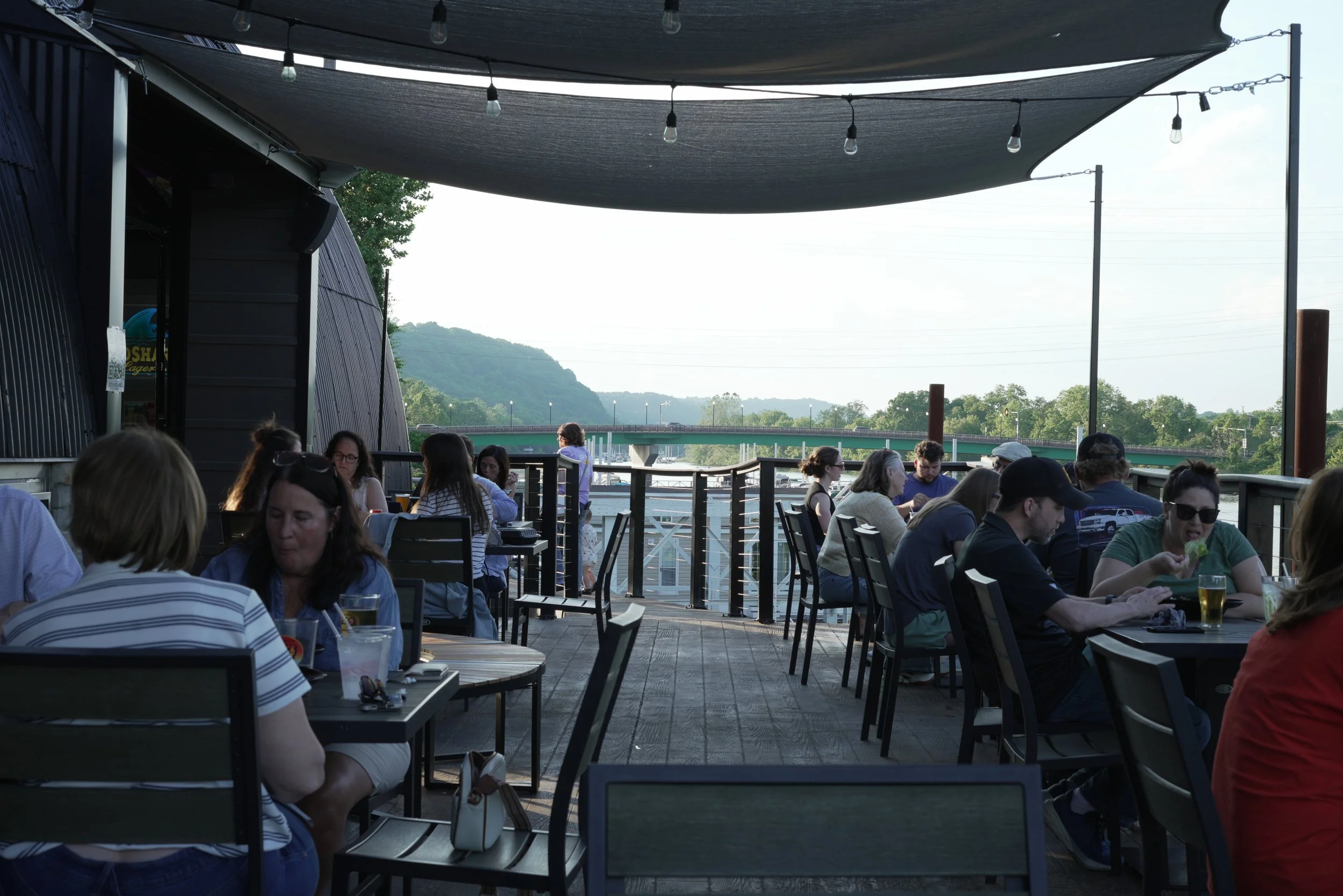People sitting at tables outdoors on a patio with a view of hills and a bridge in the background. There are shaded canopies and string lights overhead.