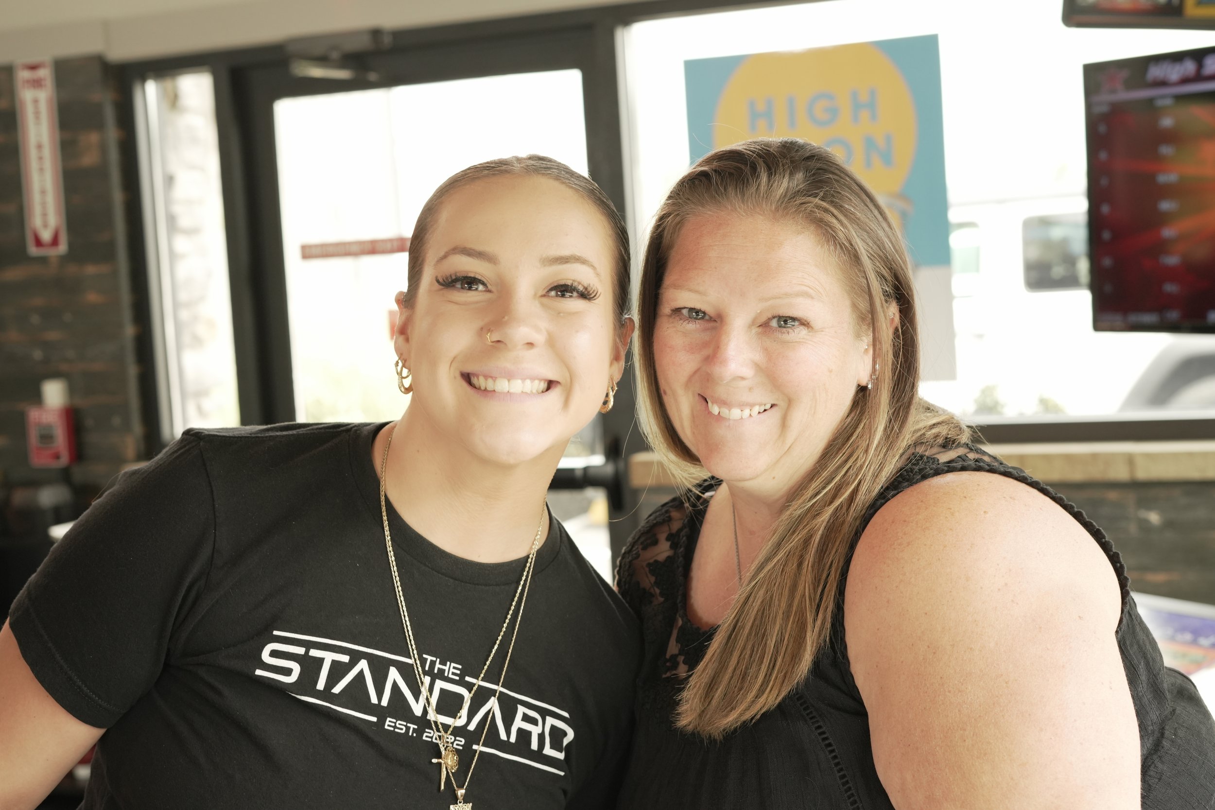 Two women smiling and taking a selfie inside a store or restaurant, with a sign that says 'HIGH ON' in the background.