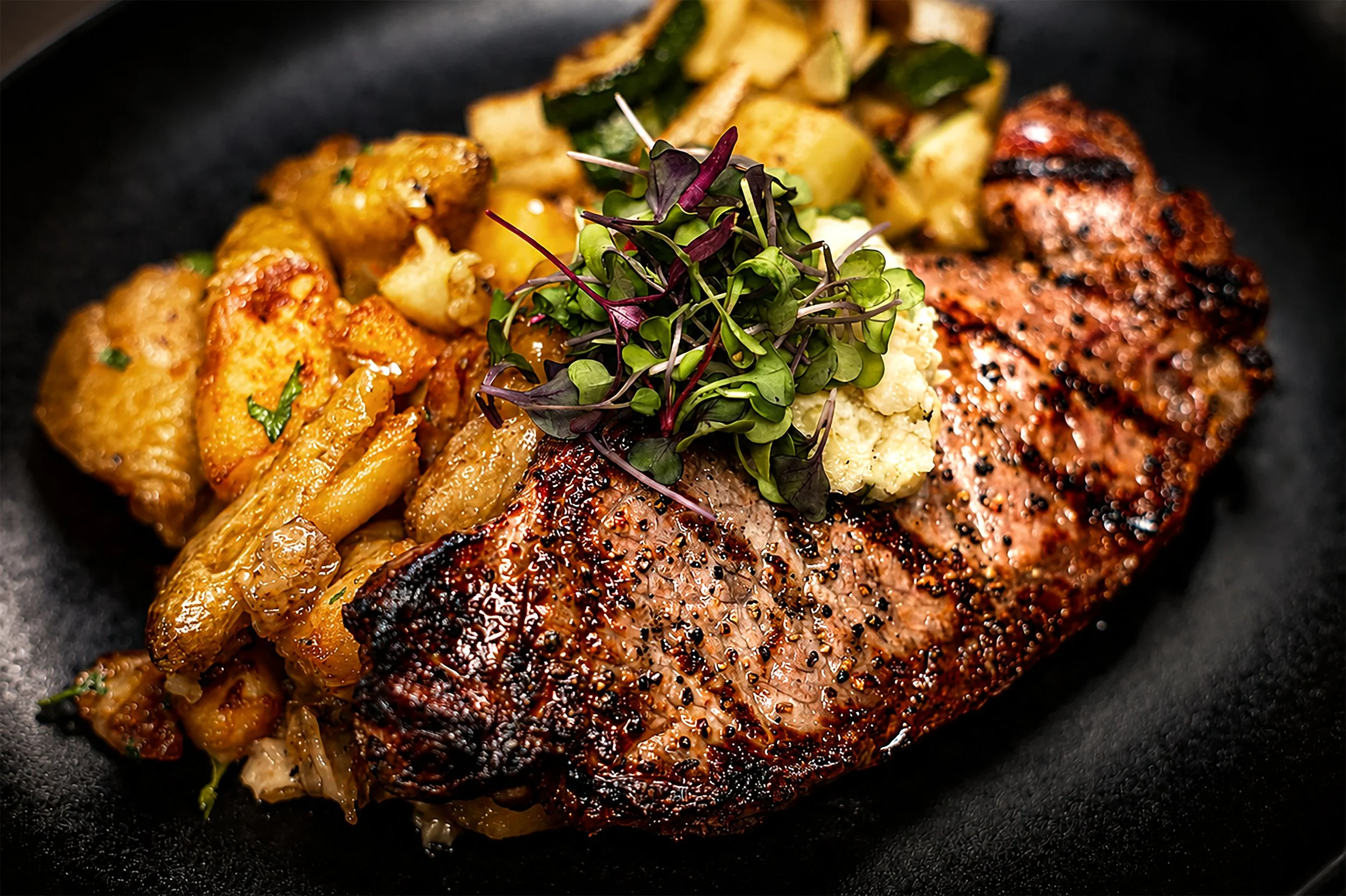 A cooked piece of seasoned salmon fillet with a black pepper crust, served with roasted cauliflower, potatoes, and sautéed zucchini, garnished with microgreens on a black plate.