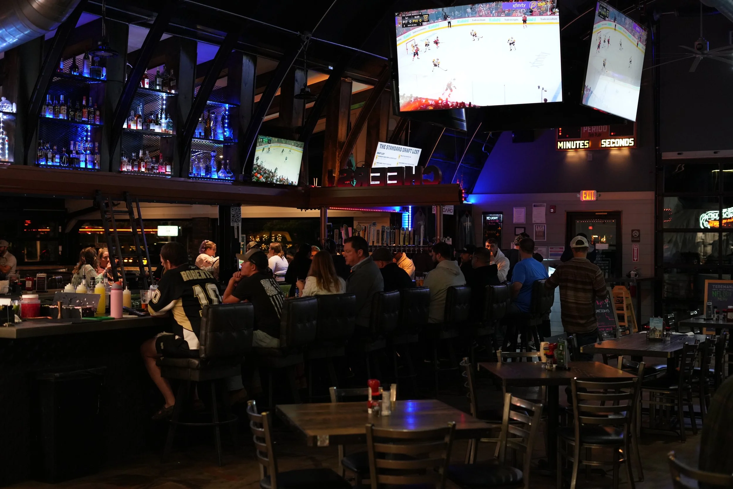 Indoor sports bar with multiple ceiling-mounted large flat-screen TVs showing a hockey game, a bar with bottles of alcohol on illuminated shelves, and patrons sitting at tables and bar stools watching the game.