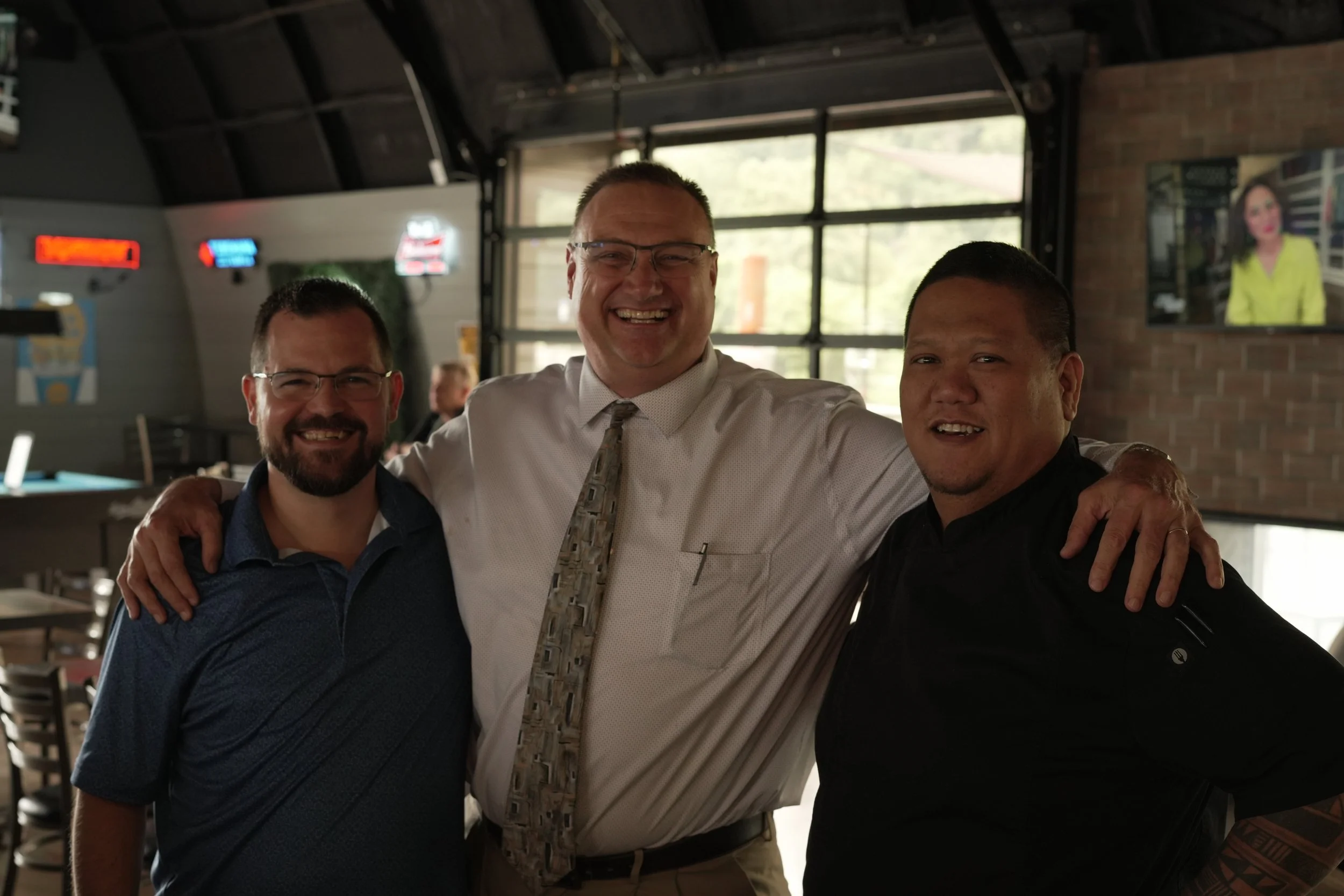 Three men smiling and posing together indoors near a window, with one man in the middle having his arms around the other two.