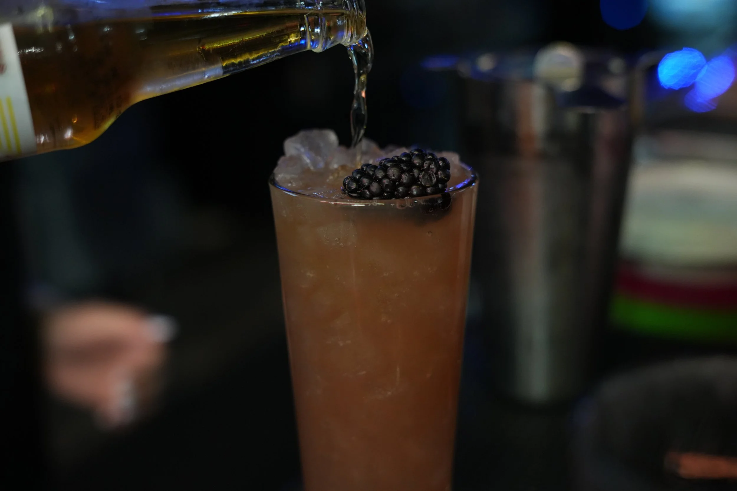 A close-up shot of a cocktail glass filled with an orange-colored drink, garnished with a cluster of blackberries on top, with a bottle pouring more liquid into the glass.
