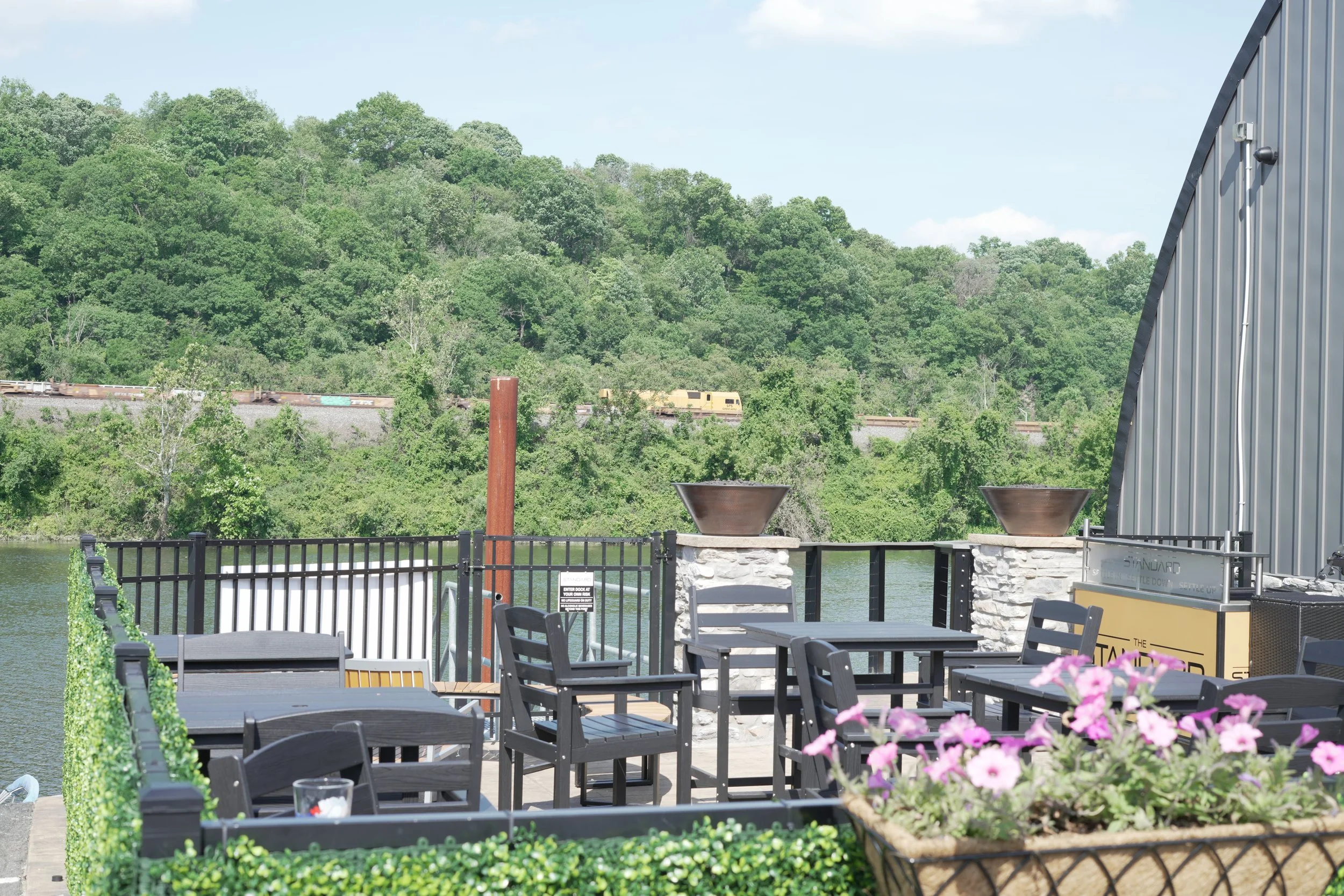 Outdoor dining setup with black tables and chairs near a body of water, with pink flowers in the foreground, a metal wall on the right, and green trees in the background.