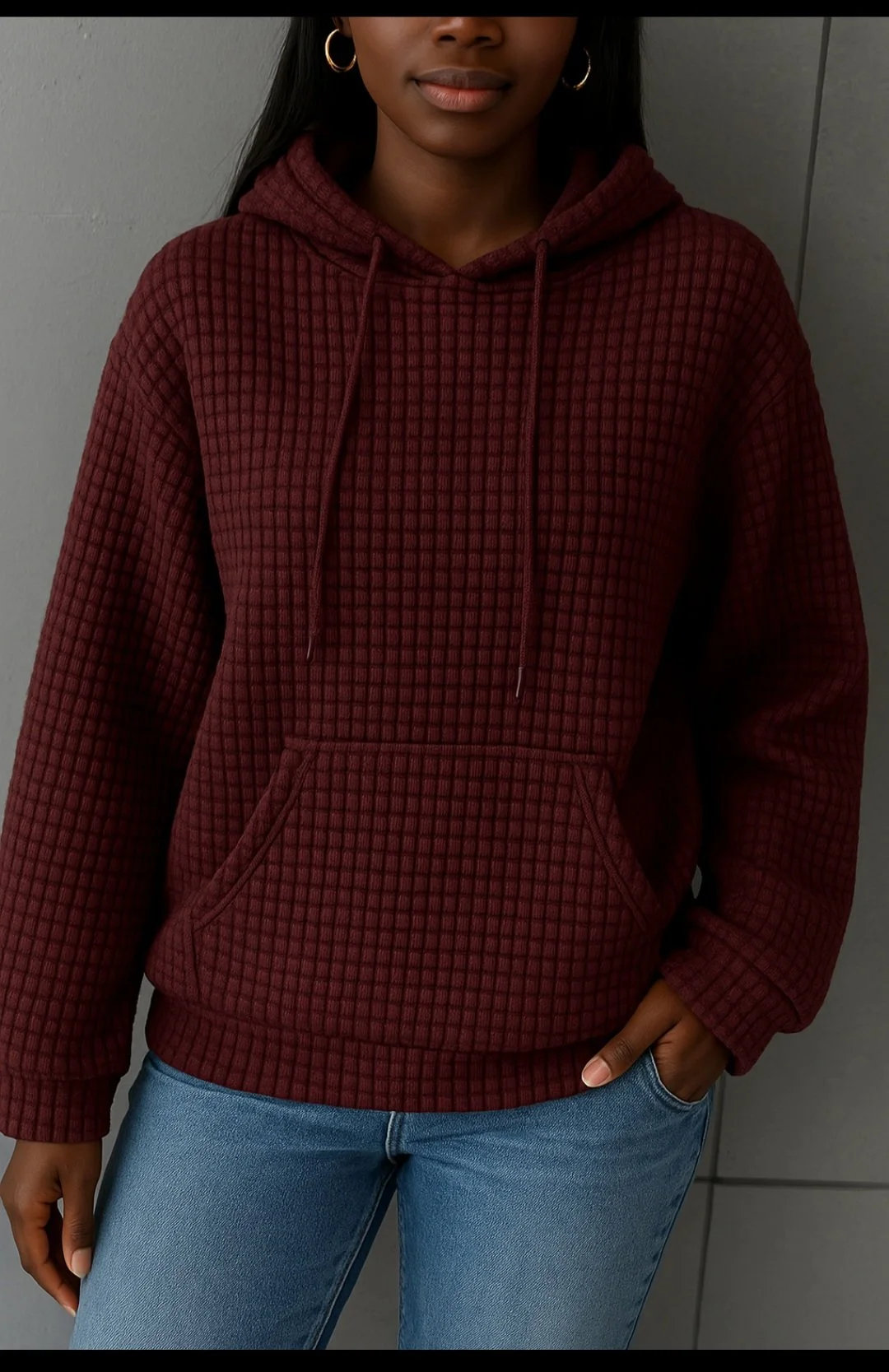 A person wearing a maroon checkered hoodie and blue jeans, with gold hoop earrings and one hand in their pocket, standing against a gray wall.