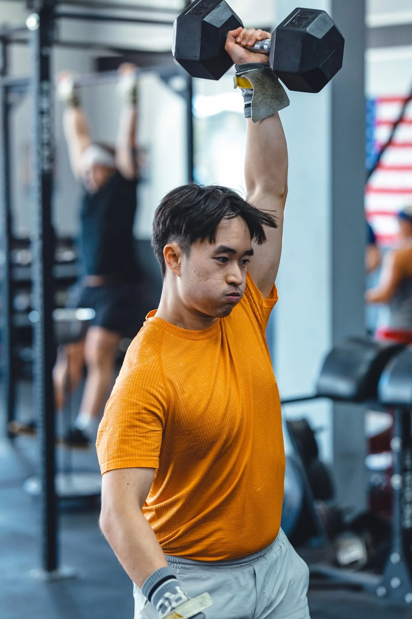 A young man lifting a black dumbbell in a gym, wearing an orange shirt and gray shorts, with other gym members and equipment visible in the background.
