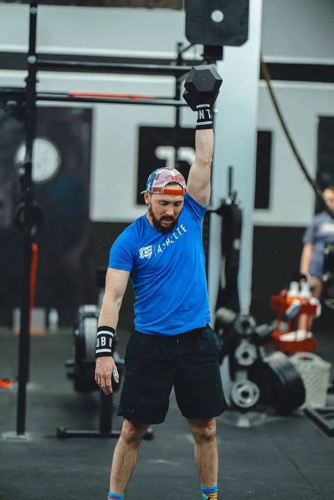 A man in a blue t-shirt and black shorts lifting a dumbbell overhead in a gym.