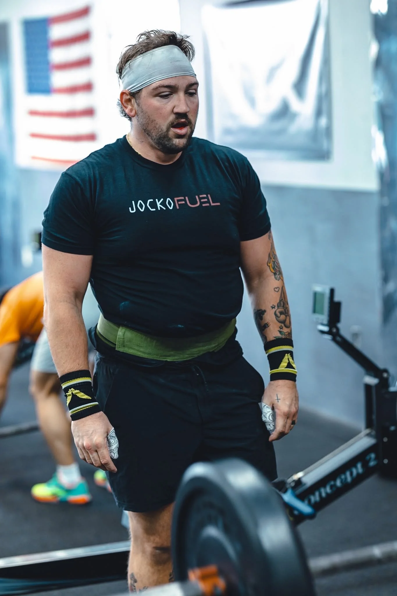A man with a beard and tattoos, wearing a gray headband, a black T-shirt with 'JOCKO FUEL' written on it, and black shorts, is working out in a gym. He appears to be exhausted after lifting weights, with a focused expression on his face. In the background, there is an American flag and gym equipment.