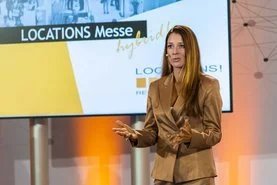Eine Frau hält eine Präsentation vor einem Bildschirm mit dem Text 'LOCATIONS Messe' auf einer Messebühne.