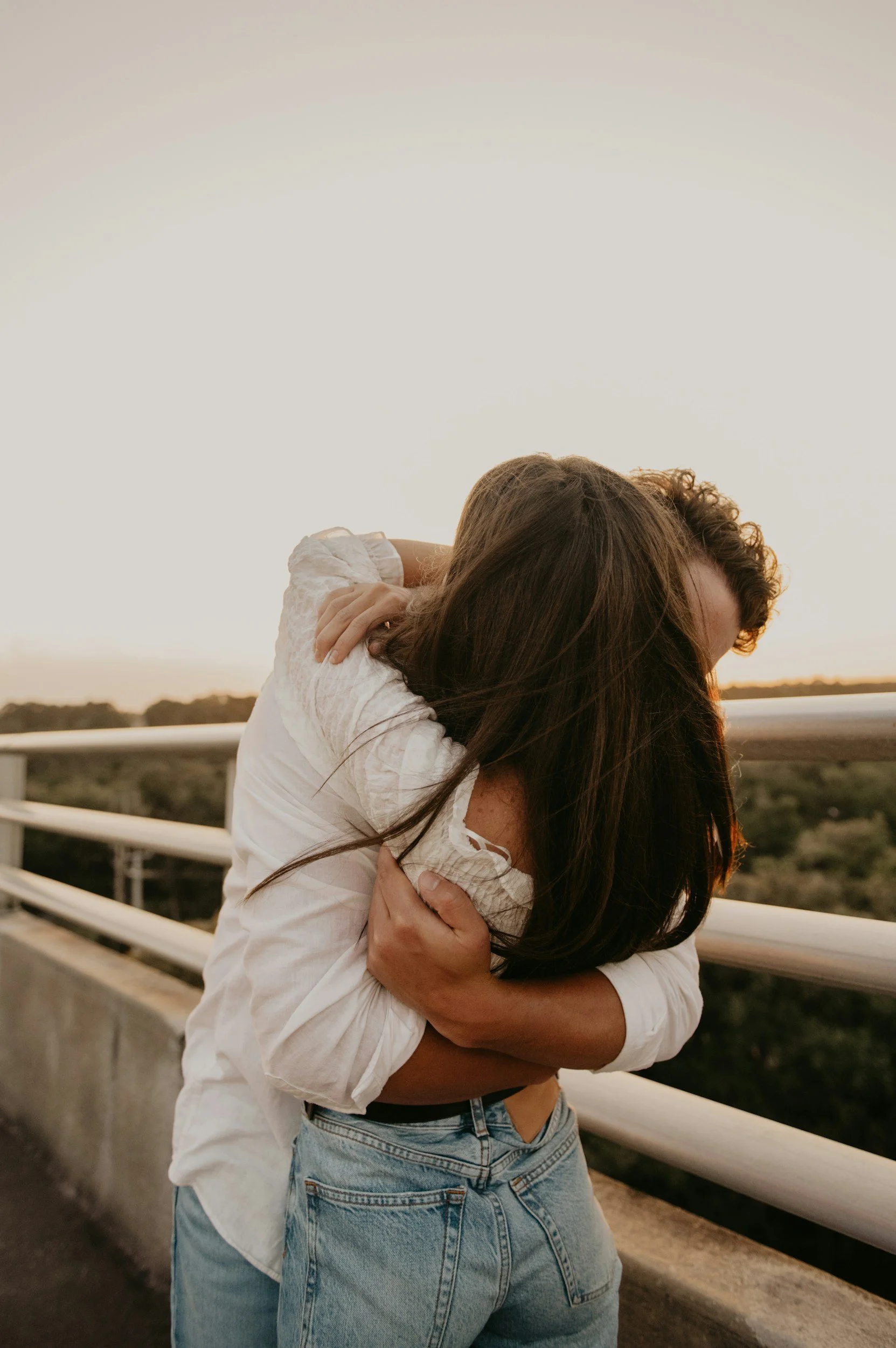 A couple hugging on a bridge during sunset, with the woman wearing a white shirt and the man in a dark shirt and jeans, embracing passionately.