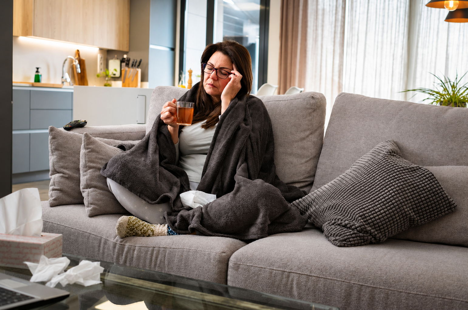 A woman sitting on a beige sofa, wrapped in a dark blanket, holding a cup of tea, with a tired or sick facial expression, in a modern living room with large windows and curtains, tissue box, and scattered tissues on a glass coffee table.