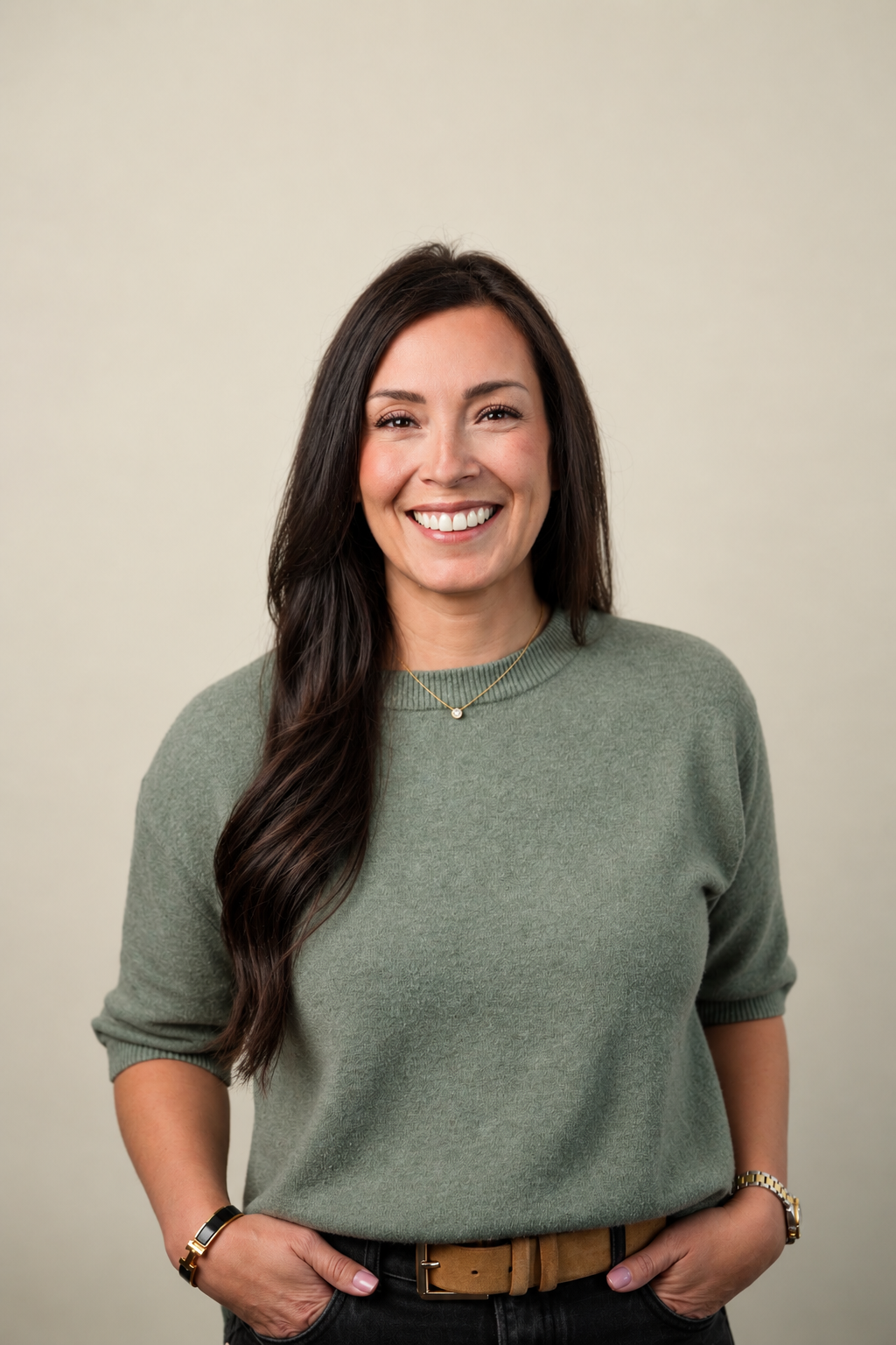 A woman with long dark hair smiling, wearing a green sweater, gold watch, bracelet, and necklace, standing against a plain light background.