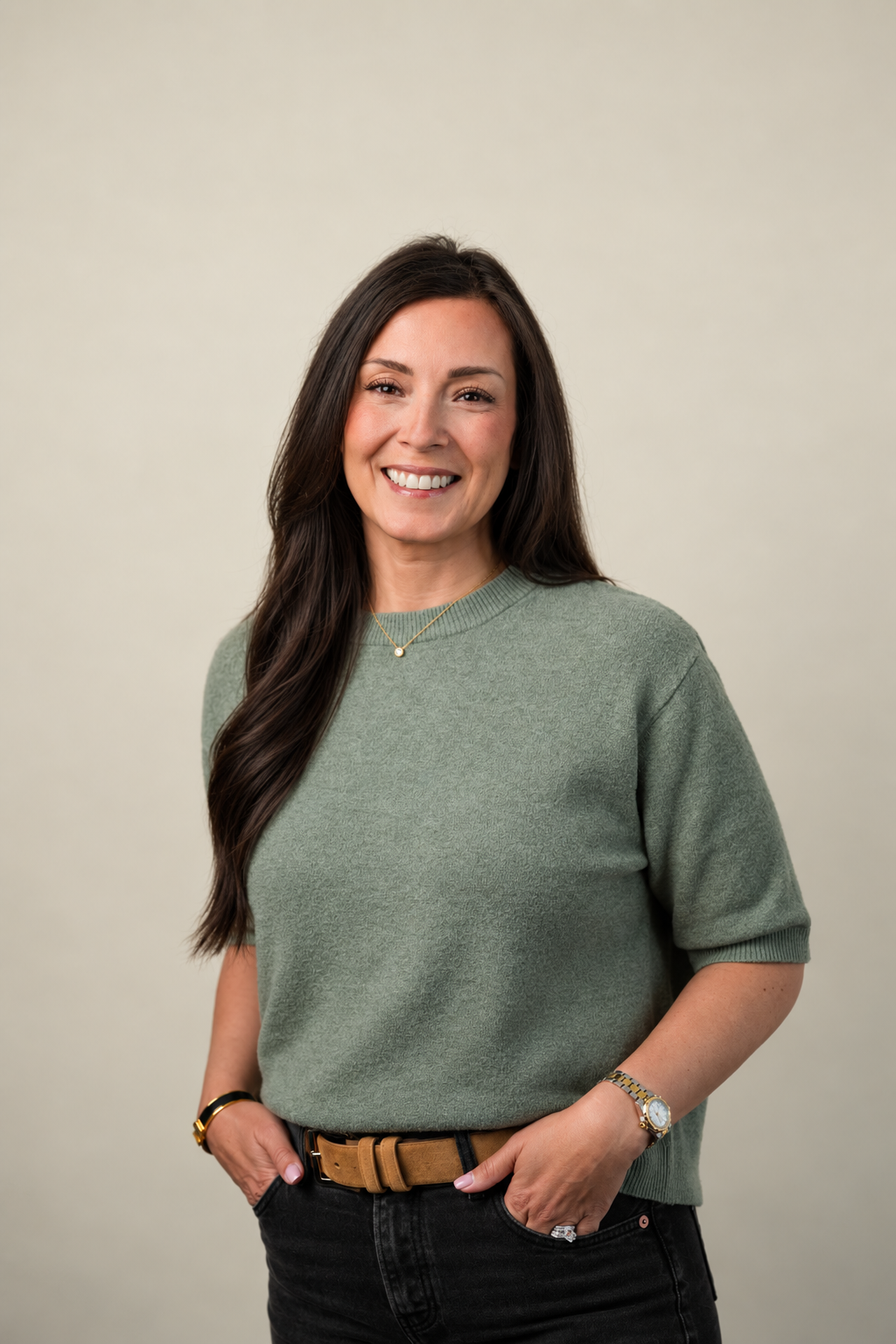 Woman with long dark hair smiling, wearing a green sweater, black pants, a gold watch, necklace, and bracelets, standing against a plain light background.