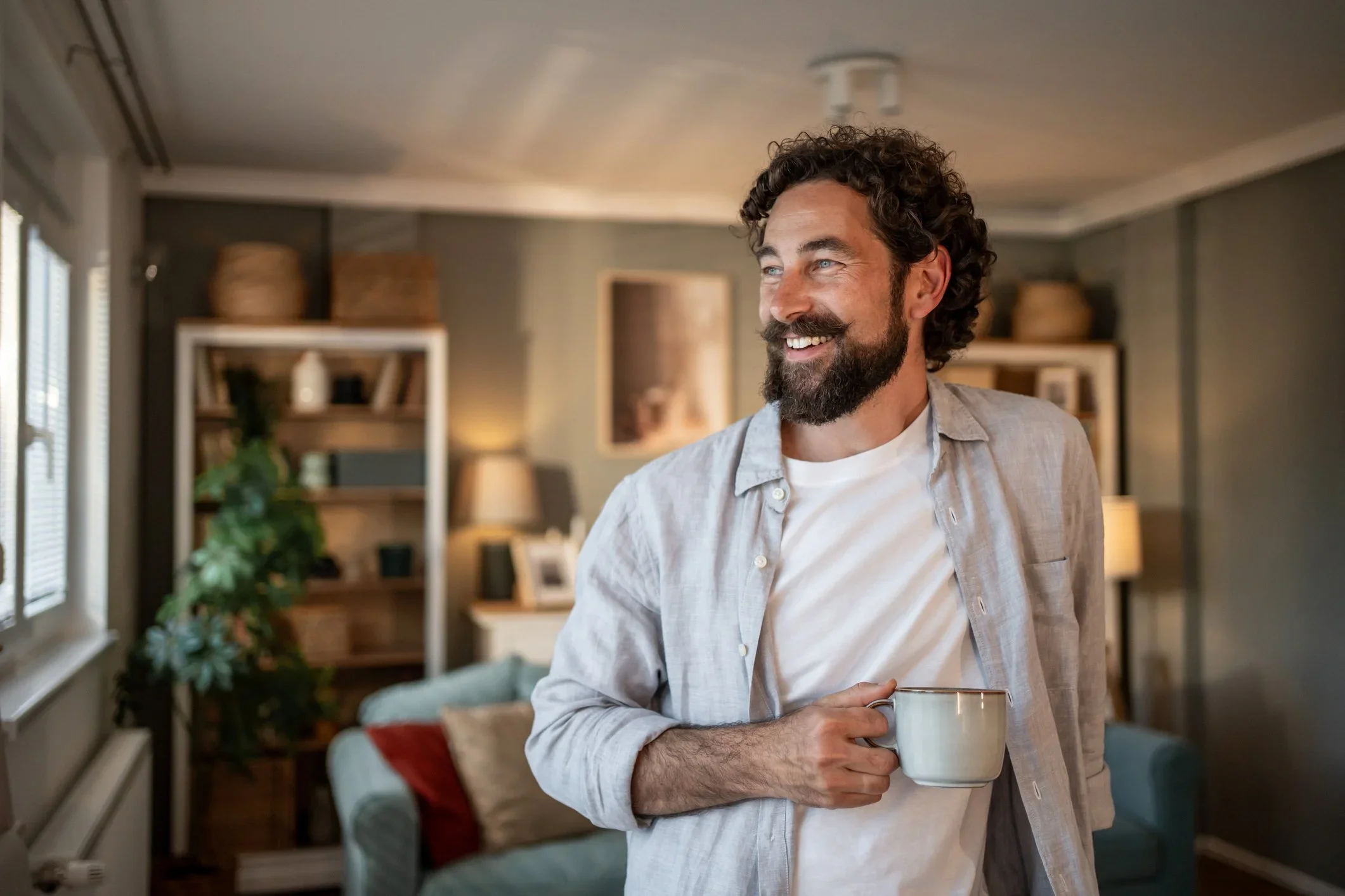 A man with curly hair, beard, and mustache smiling and holding a mug in a cozy living room.