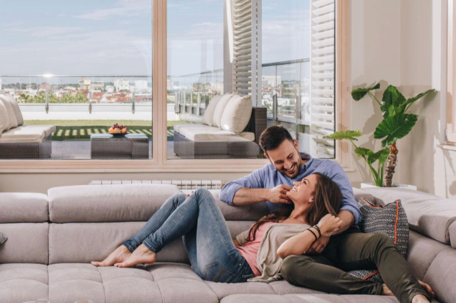 Smiling couple relaxing on a light-colored sofa in a bright living room with large windows showing an outdoor balcony and city view, with houseplants nearby.