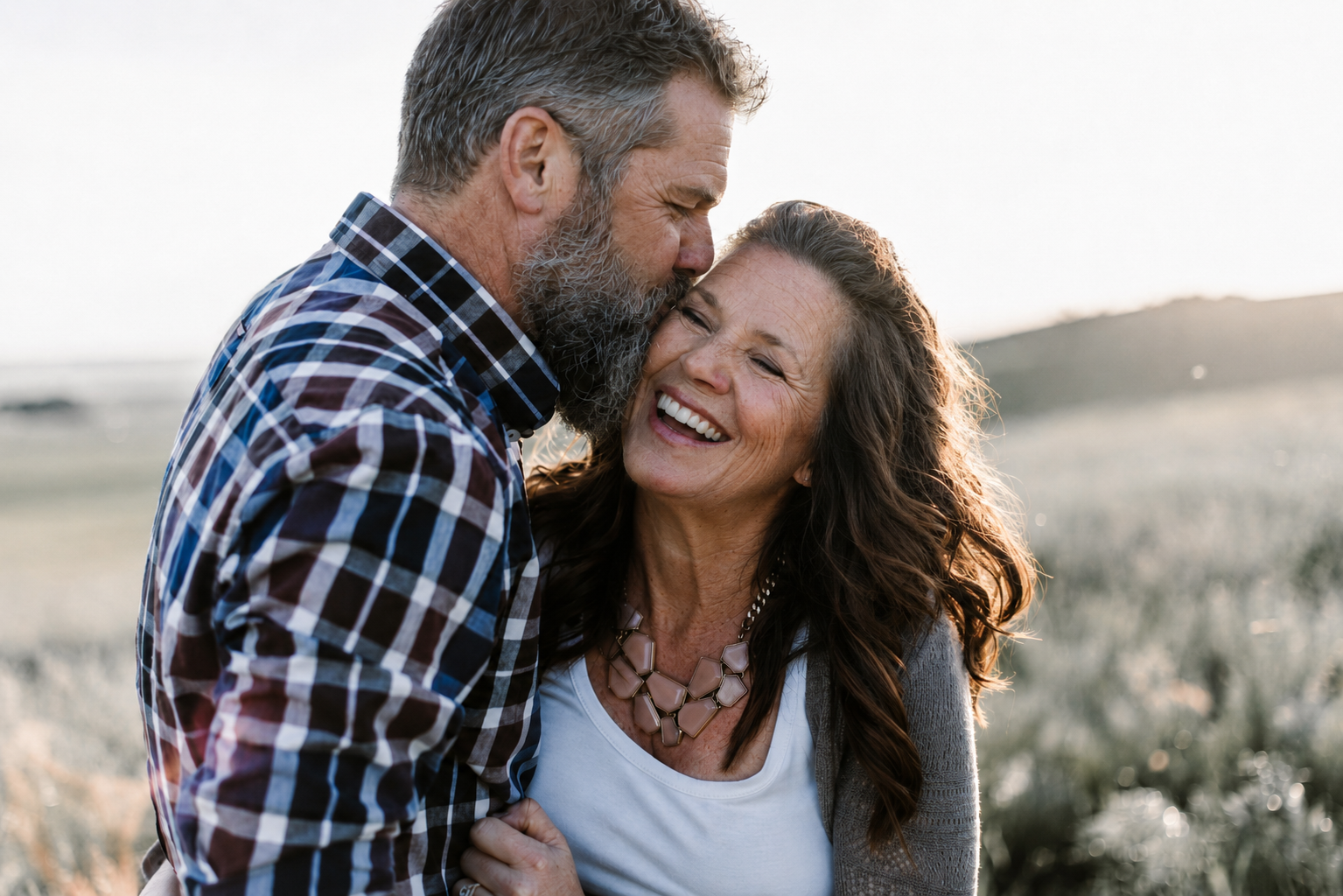 An elderly couple sharing a joyful moment outdoors at sunset, with the man kissing the woman's forehead and the woman smiling happily.