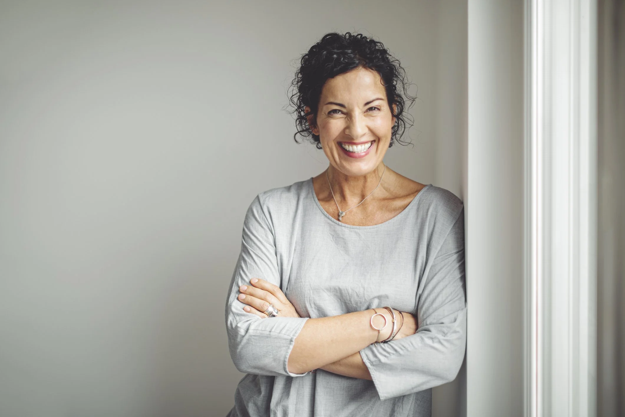 A smiling woman with short curly black hair, wearing a light gray long-sleeve shirt, standing with arms crossed by a window, in a room with plain light-colored walls.