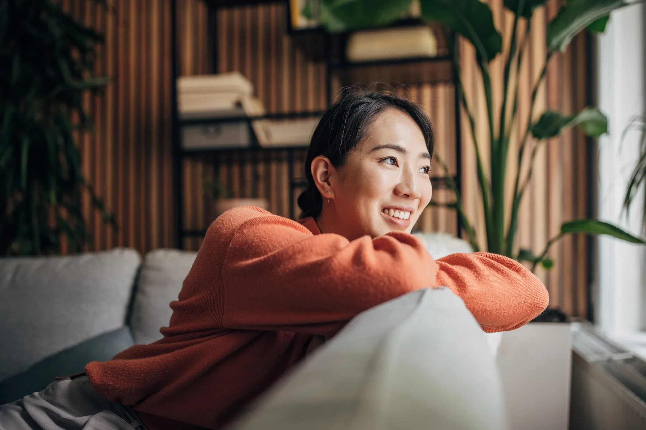 A young woman with dark hair and light skin smiling and leaning on her arms, sitting on a light-colored sofa in a cozy room with wooden wall panels and green plants.