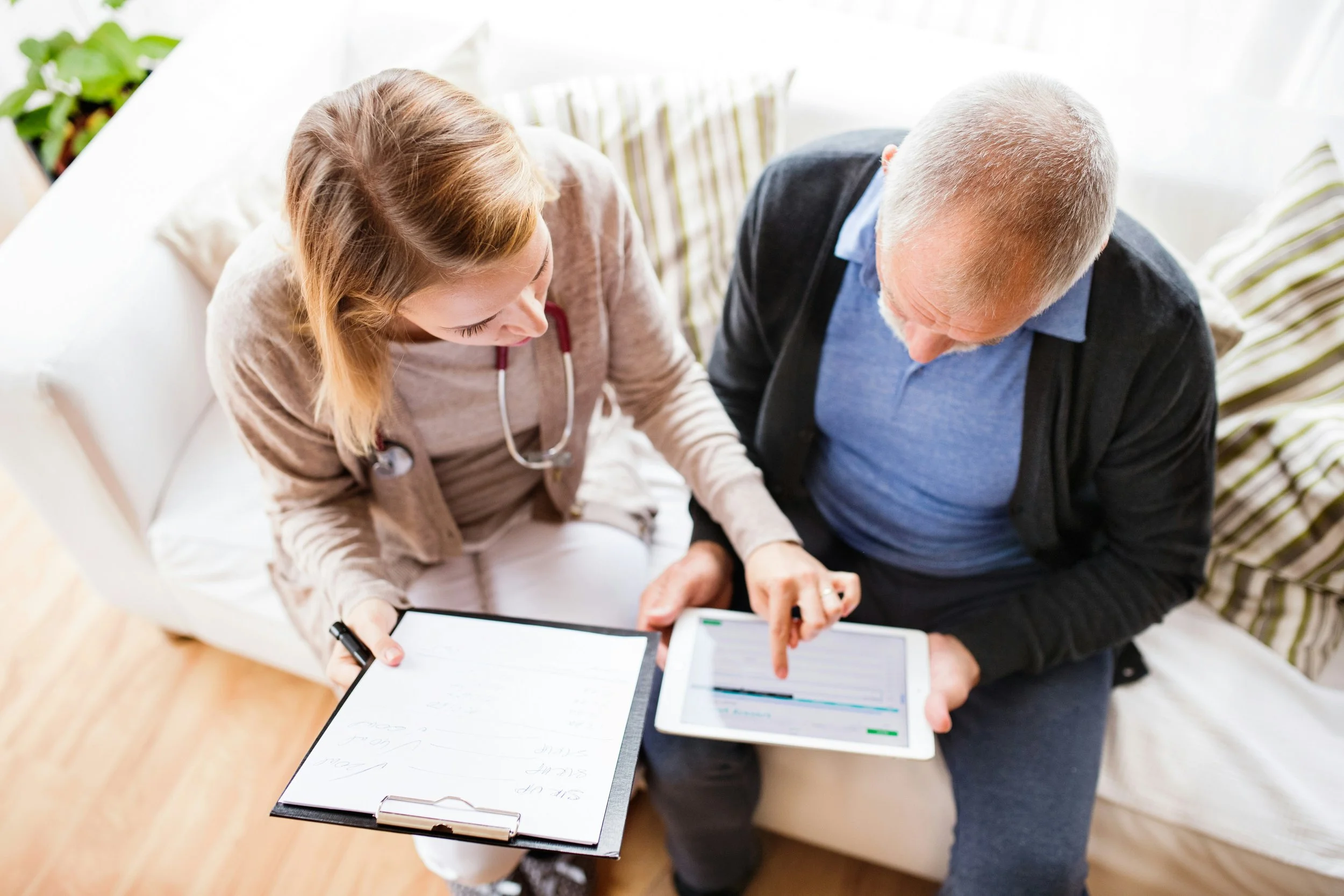 A private nurse advocate and an elderly male client are sitting together on a couch during a home visit, looking at medical documents and a tablet, in a bright room with natural light in the client's home in Raleigh, NC.