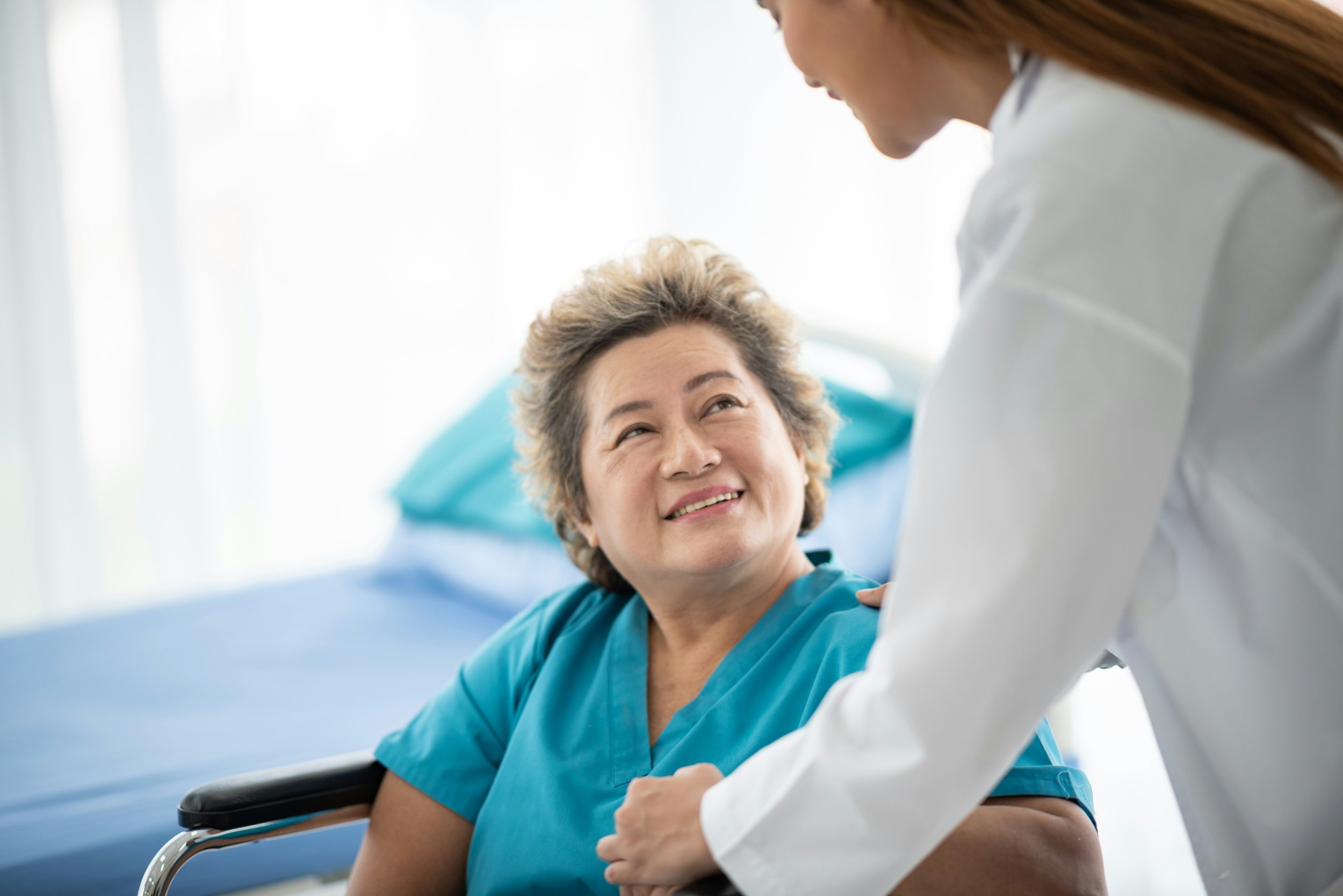 A nurse advocate is smiling at an elderly woman in a hospital room, engaging in a caring conversation to help communication between medical providers, family, and the client. The nurse advocate will help make sure the client's voice is heard.