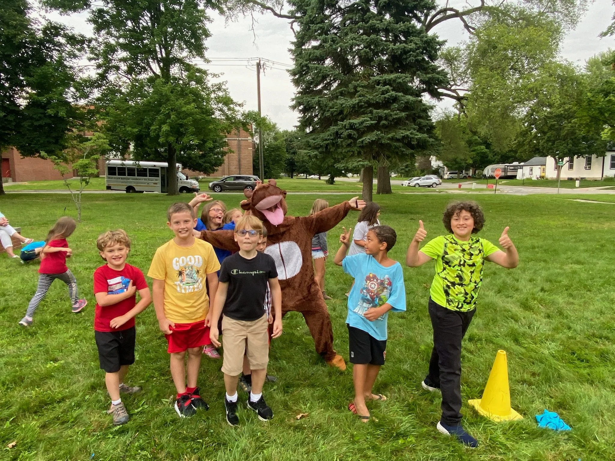 A group of children smiling and posing for the camera outdoors on a grassy area with trees in the background. There is a person in a bear costume among them. Some children are giving thumbs-up signs.