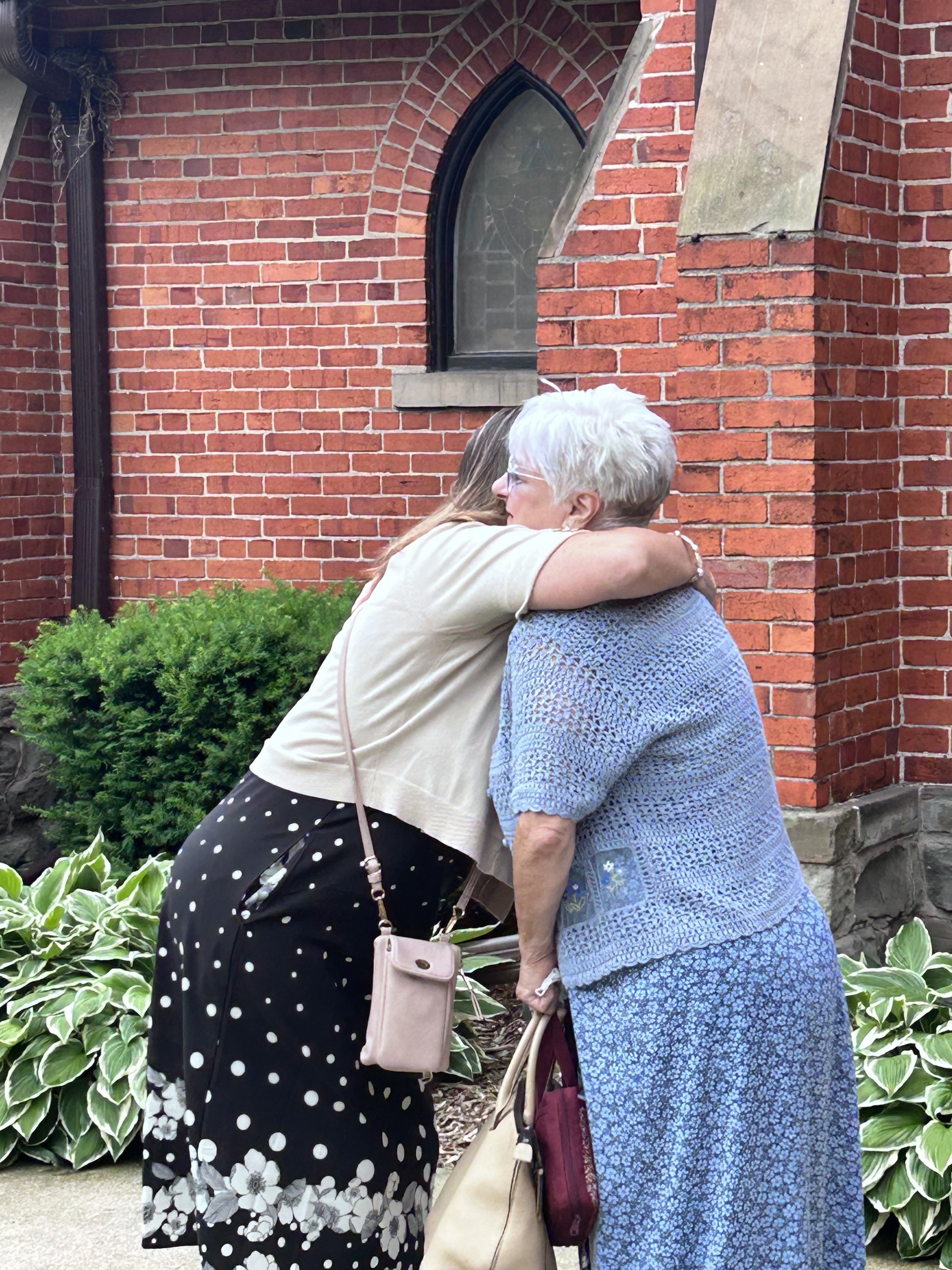 An elderly woman and a younger woman hugging in front of a brick building with arched window and green plants.