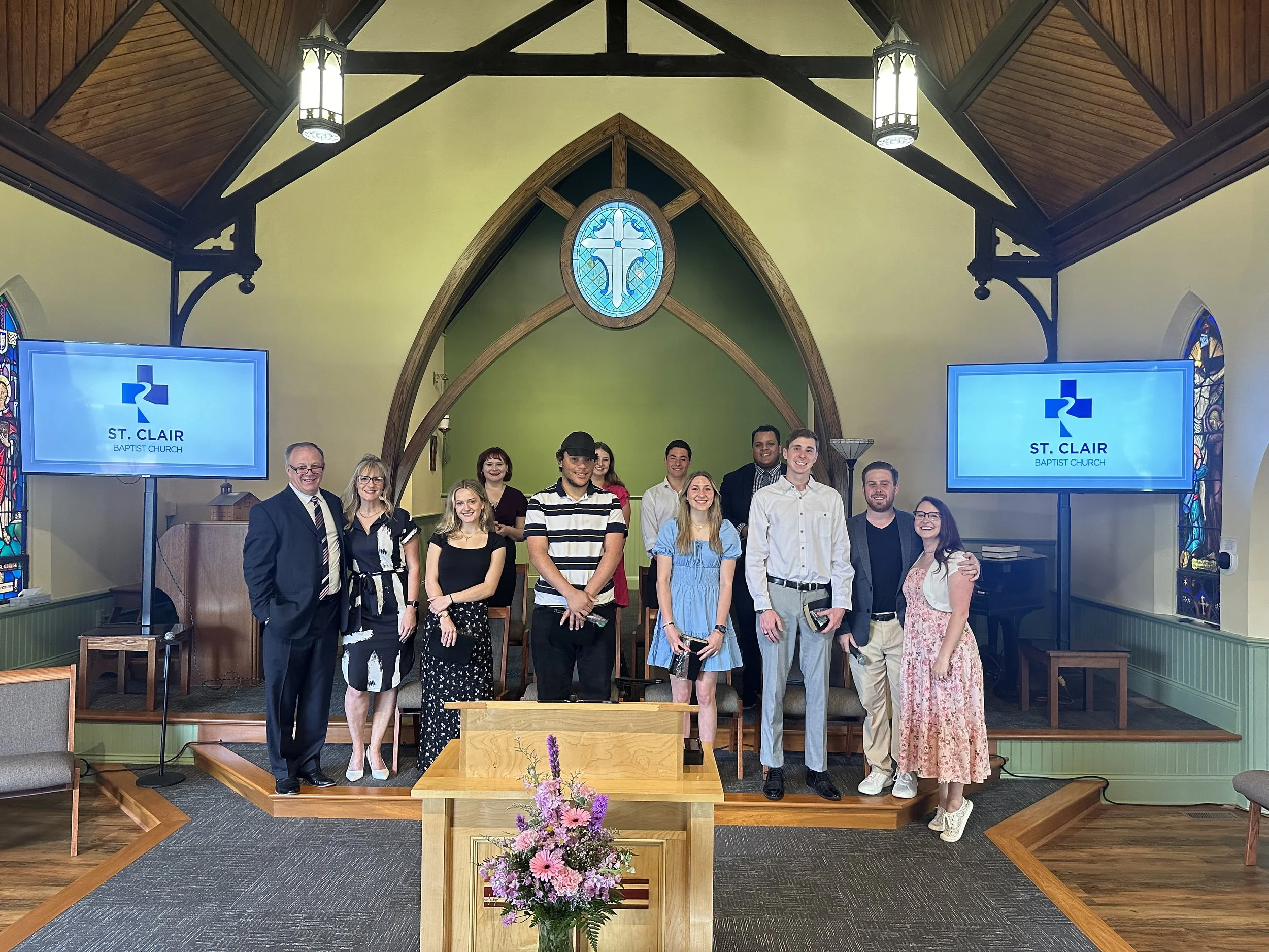 Group of people standing on stage inside a church, with a wooden cross and stained glass windows, for a church event or ceremony at St. Clair Baptist Church.