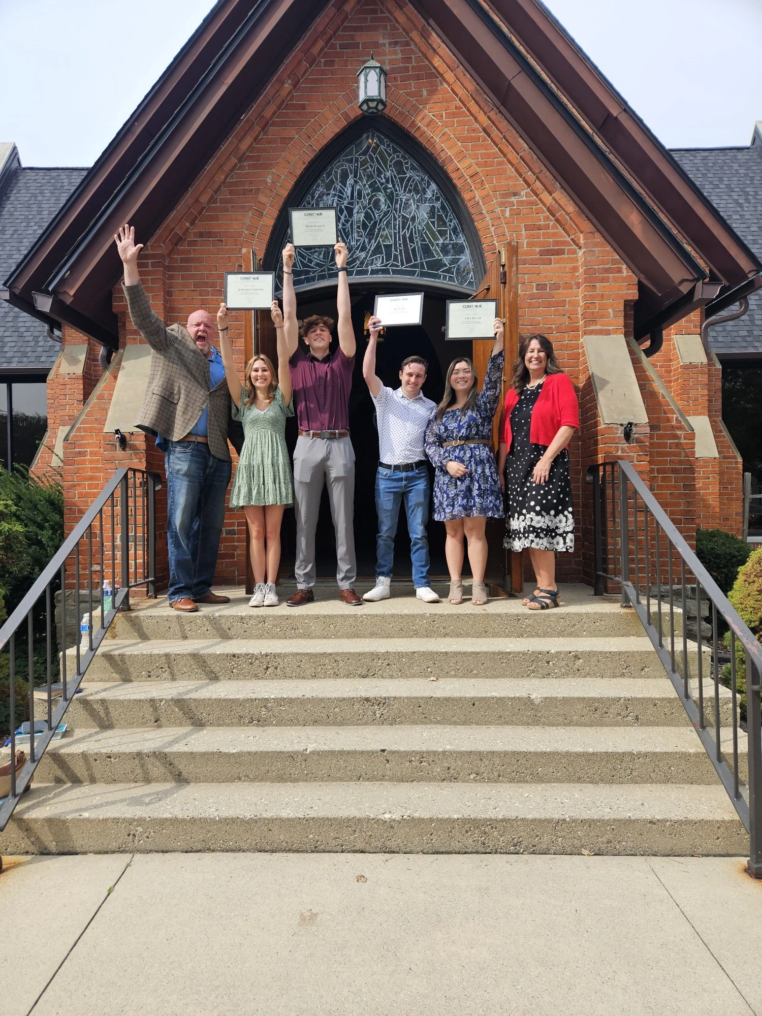 Group of six people standing on the steps of a brick church, celebrating and holding up certificates, with one person raising an arm in excitement.