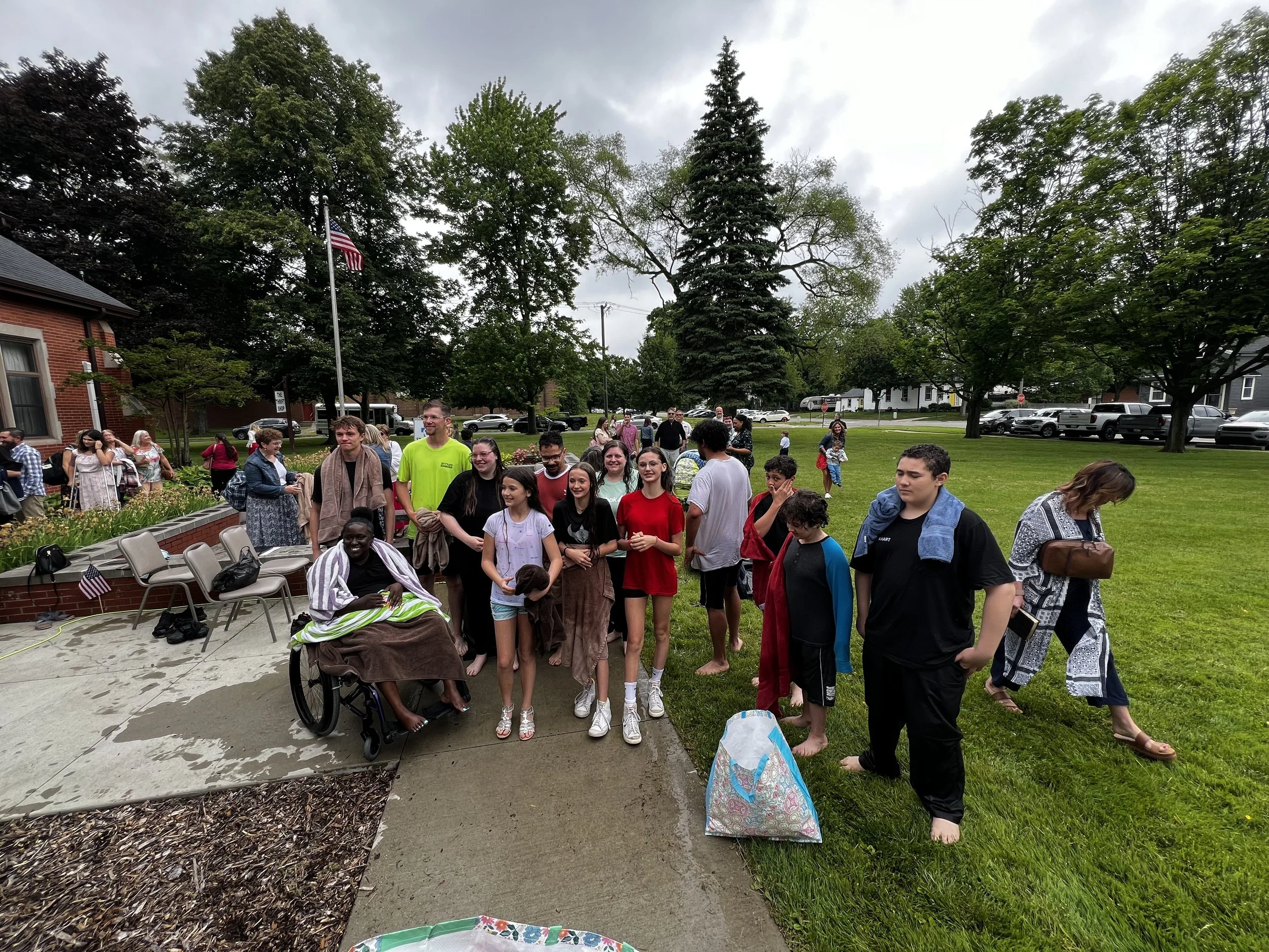 A group of people gathered outdoors in a park-like setting with trees and a cloudy sky, some in casual attire and some in dressier clothing, standing on a sidewalk and grassy area, with a brick building and parked cars in the background.