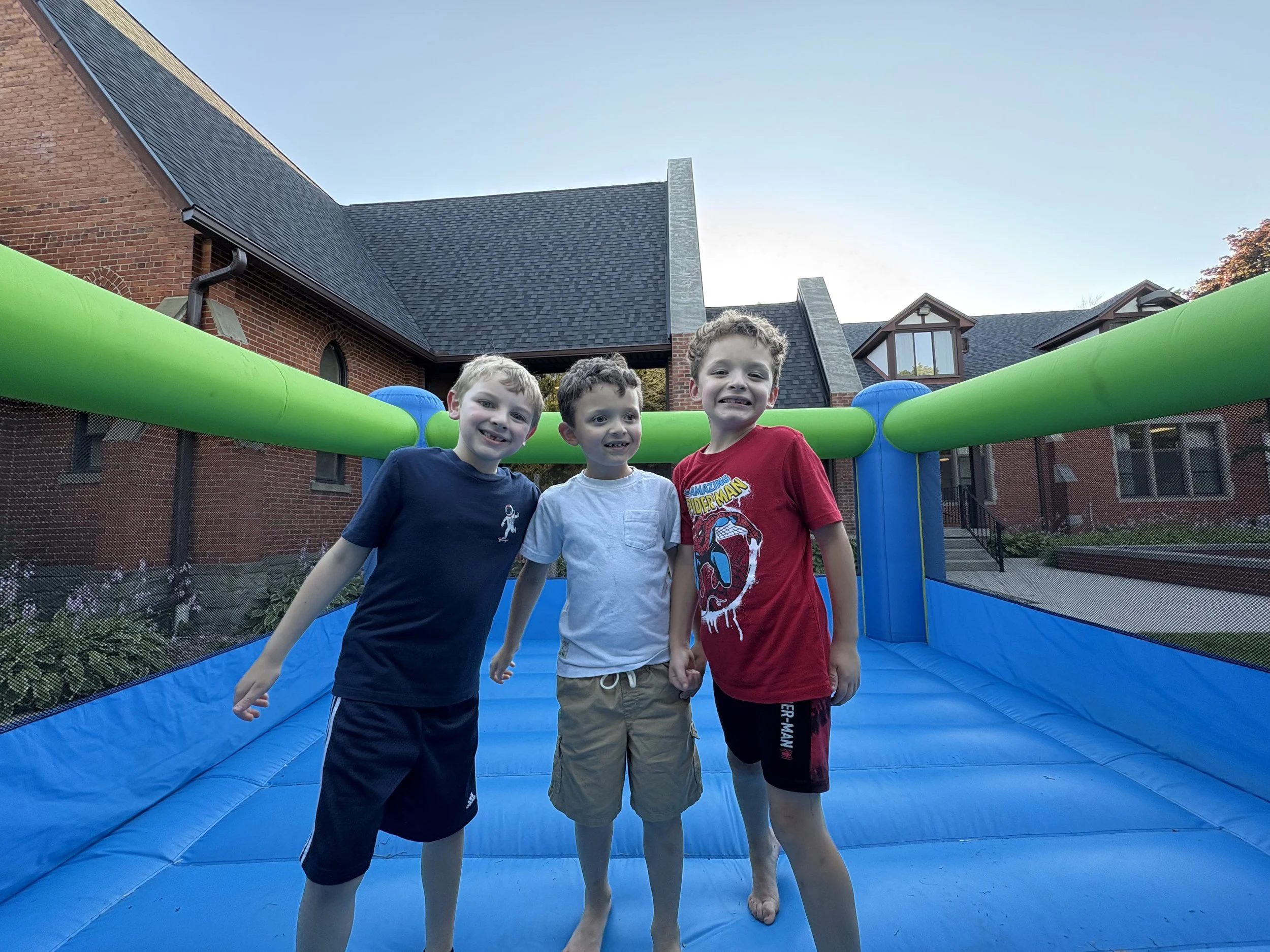 Three young boys standing on an inflatable bounce house, smiling at the camera, with a brick house and blue sky in the background.