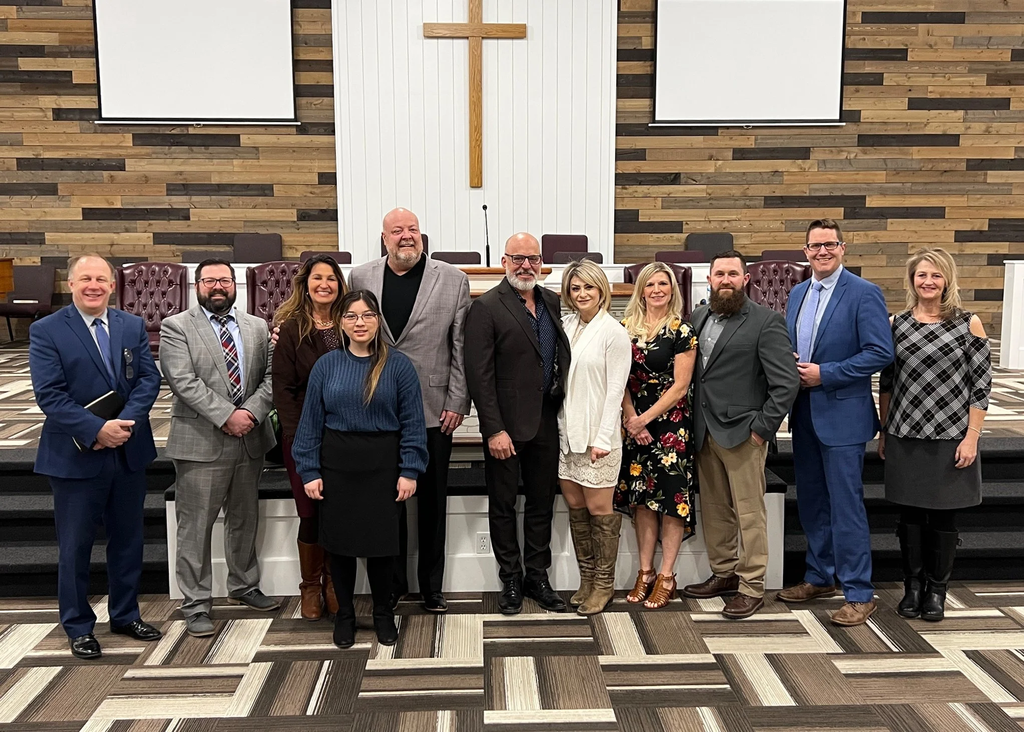A group of twelve people standing inside a church with wood-paneled walls and a wooden cross behind them, all smiling for a group photo.