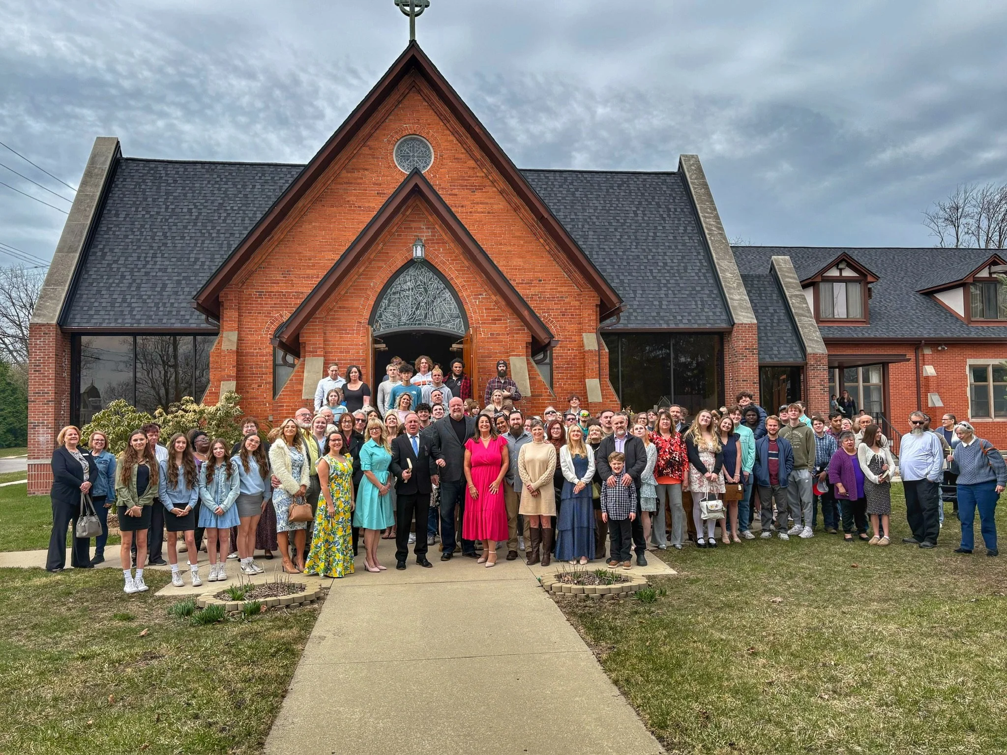 A large group of people standing in front of a red brick church with stained glass windows and a cross on top, outdoors on a cloudy day.