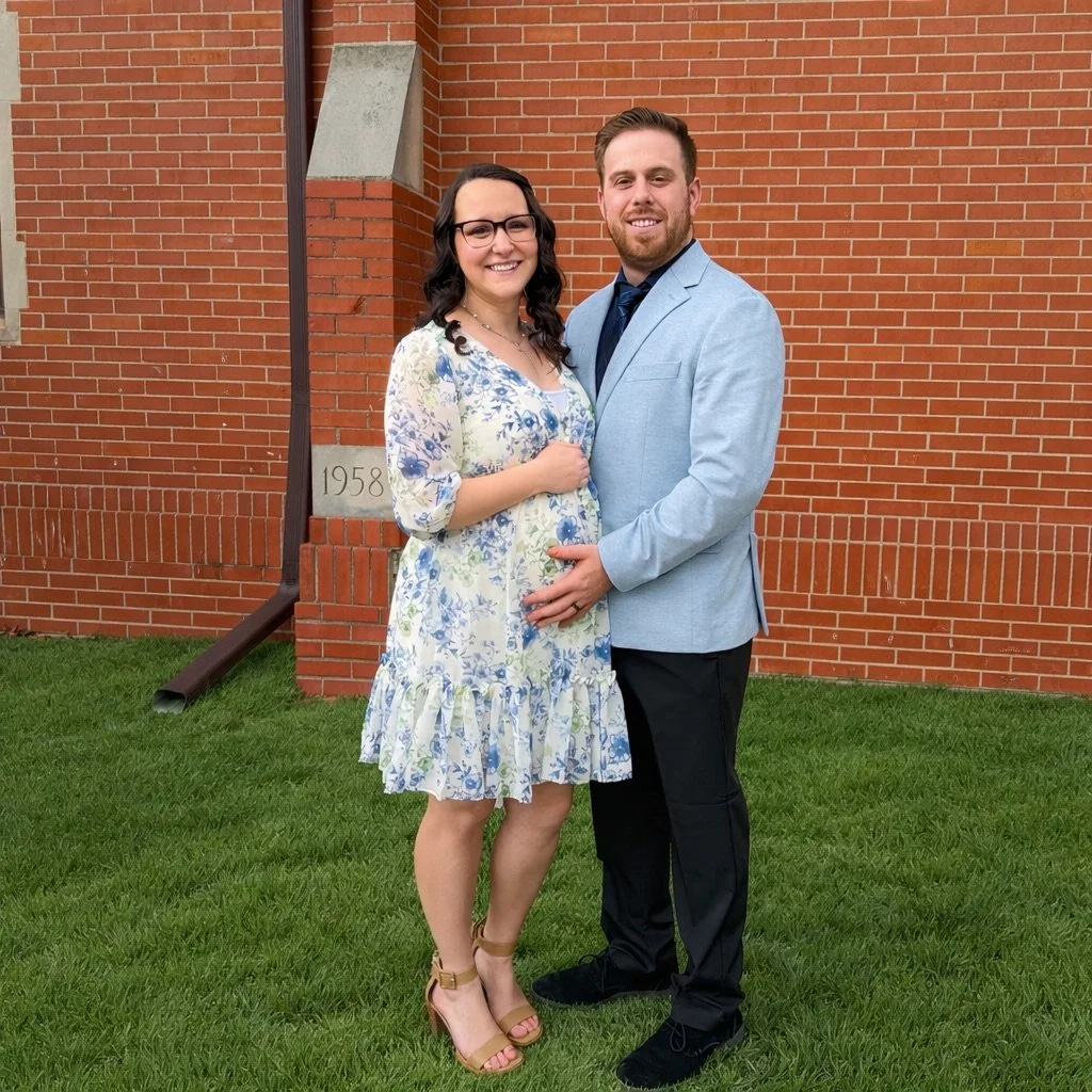 A pregnant woman and a man standing outside in front of a brick building, smiling and holding her belly, dressed in semi-formal attire.