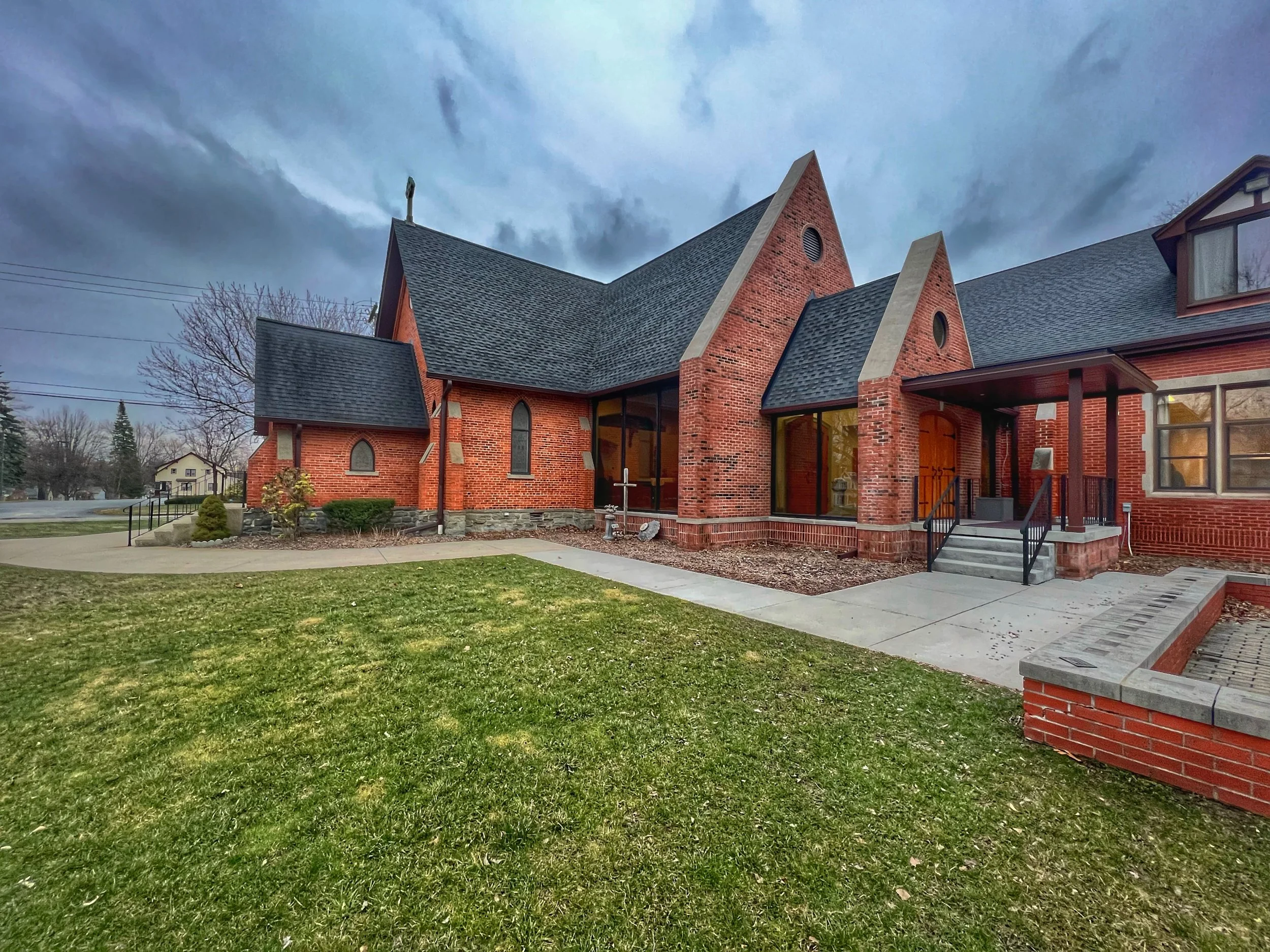 A brick church with a steep roof and pointed gables, large windows, front steps, and a cross on top, surrounded by a grassy lawn and trees under a cloudy sky.