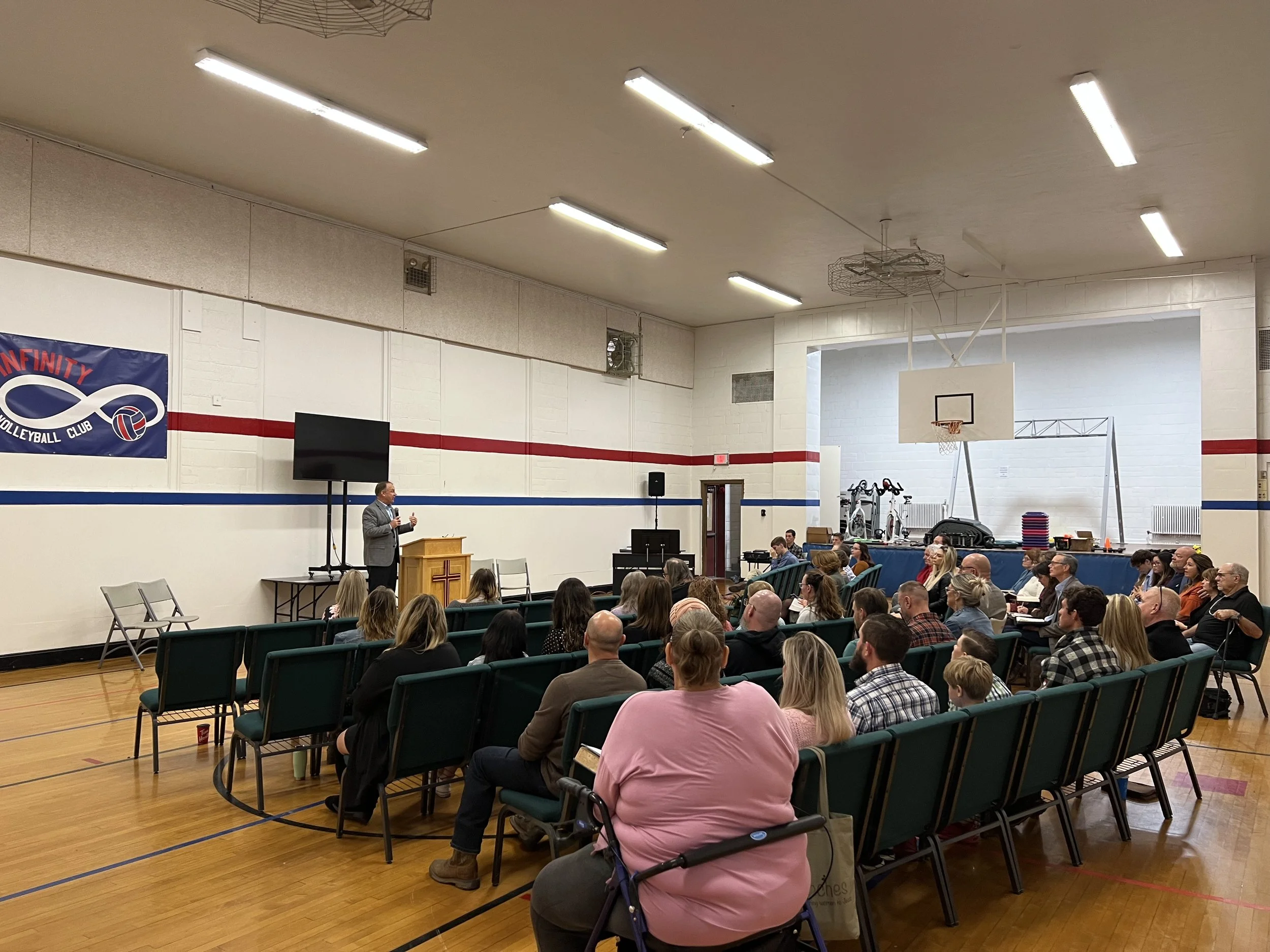 A speaker in a gray suit is giving a presentation to an audience seated in a gymnasium with a basketball hoop and a large TV screen. The room has fluorescent lighting and white walls with a volleyball club banner.