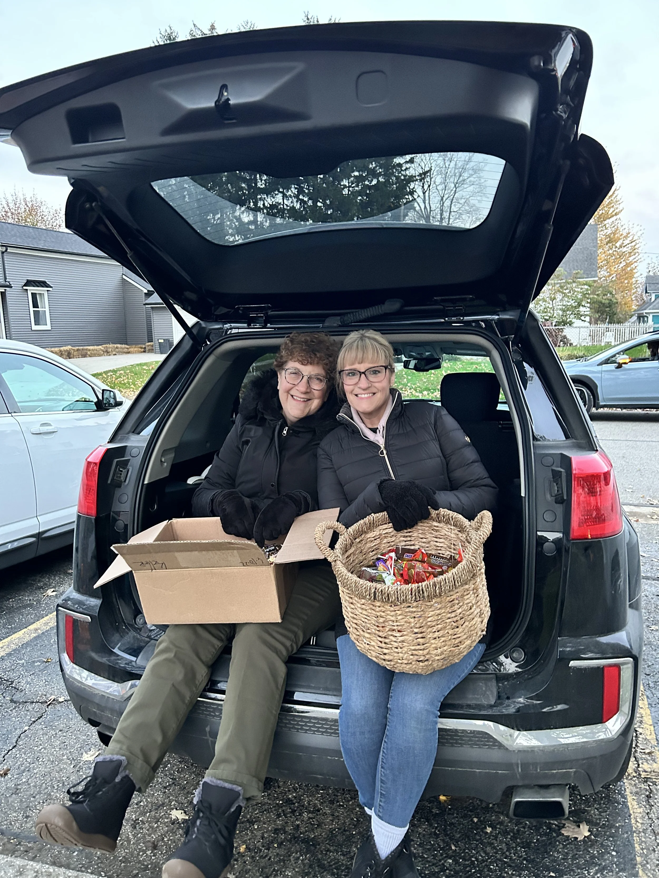 Two women sitting in the open trunk of a black vehicle, smiling, with a box and a woven basket filled with candy in front of them, in a parking lot with other cars and buildings visible in the background.