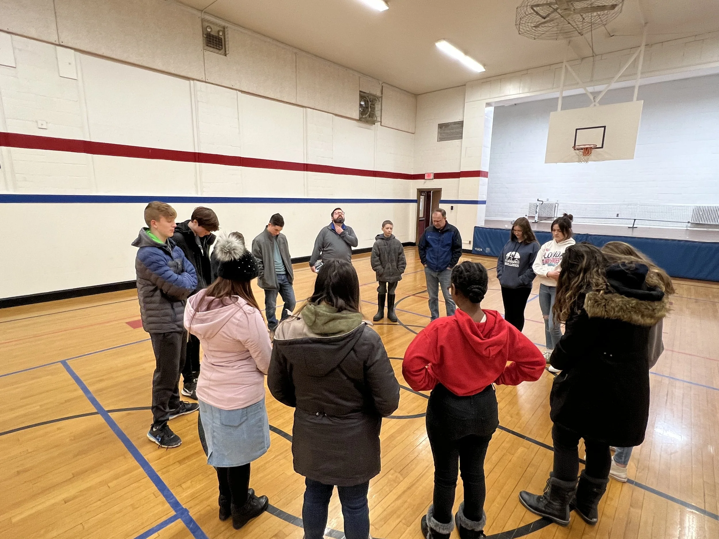 A group of people standing in a circle inside a gymnasium. Some individuals have their heads bowed while others look up. The gym has a basketball hoop and wooden flooring, with a red and blue line on the wall.