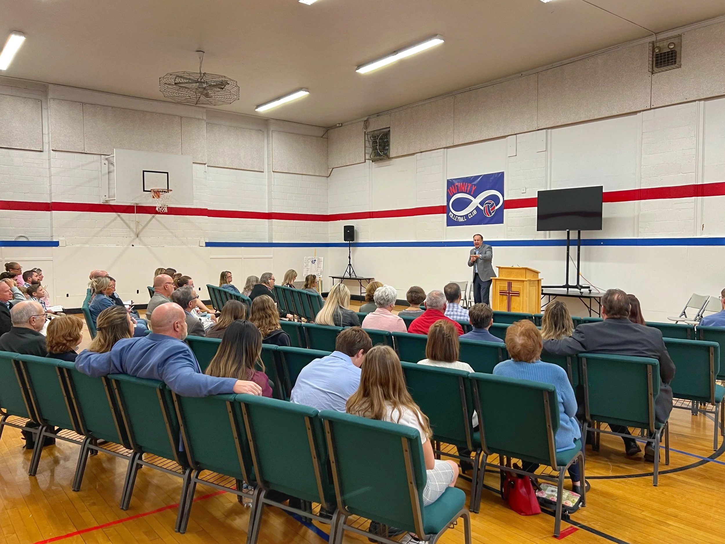 A gathering in a gymnasium with a speaker standing at a podium, addressing an audience seated in green chairs. The gym has a basketball hoop and a banner that reads 'Infinity Volleyball Club.'