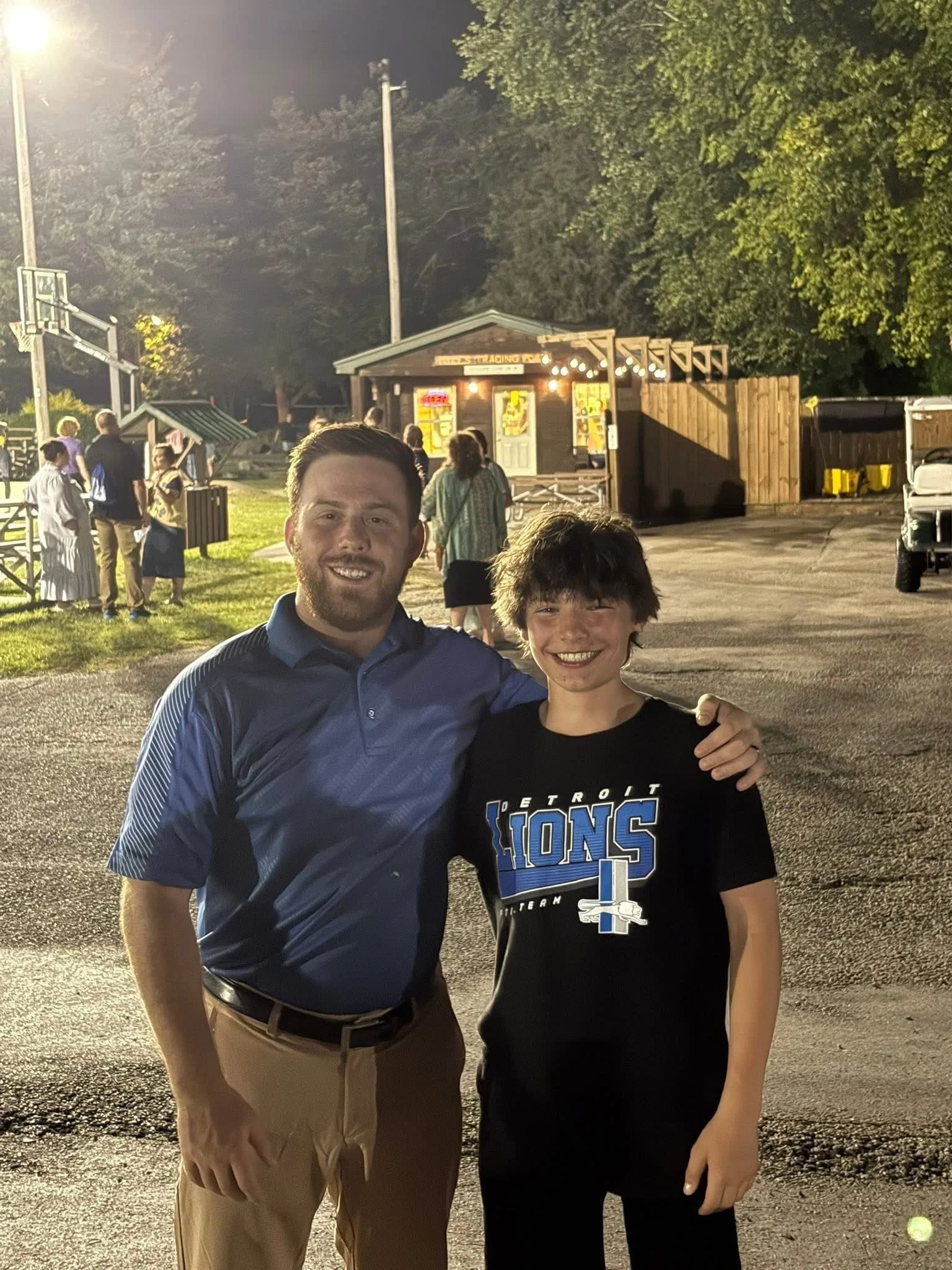 Two males, one adult and one young, standing outside at night, smiling for a selfie. Behind them, there are people in line, trees, a small building with lights, and a basketball hoop.