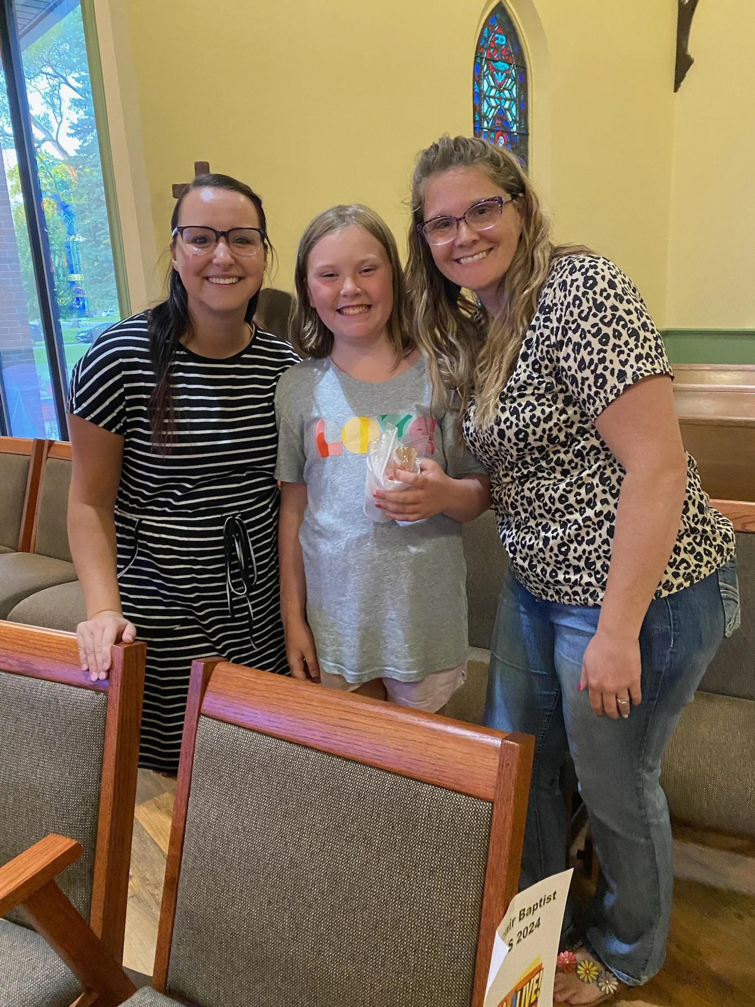 Three women and a girl smiling inside a church. The girl, in the middle, is holding a small clear bag. The women are wearing glasses; one is wearing a striped black and white shirt, and the other has a leopard print top. There is stained glass and a cross visible in the background.