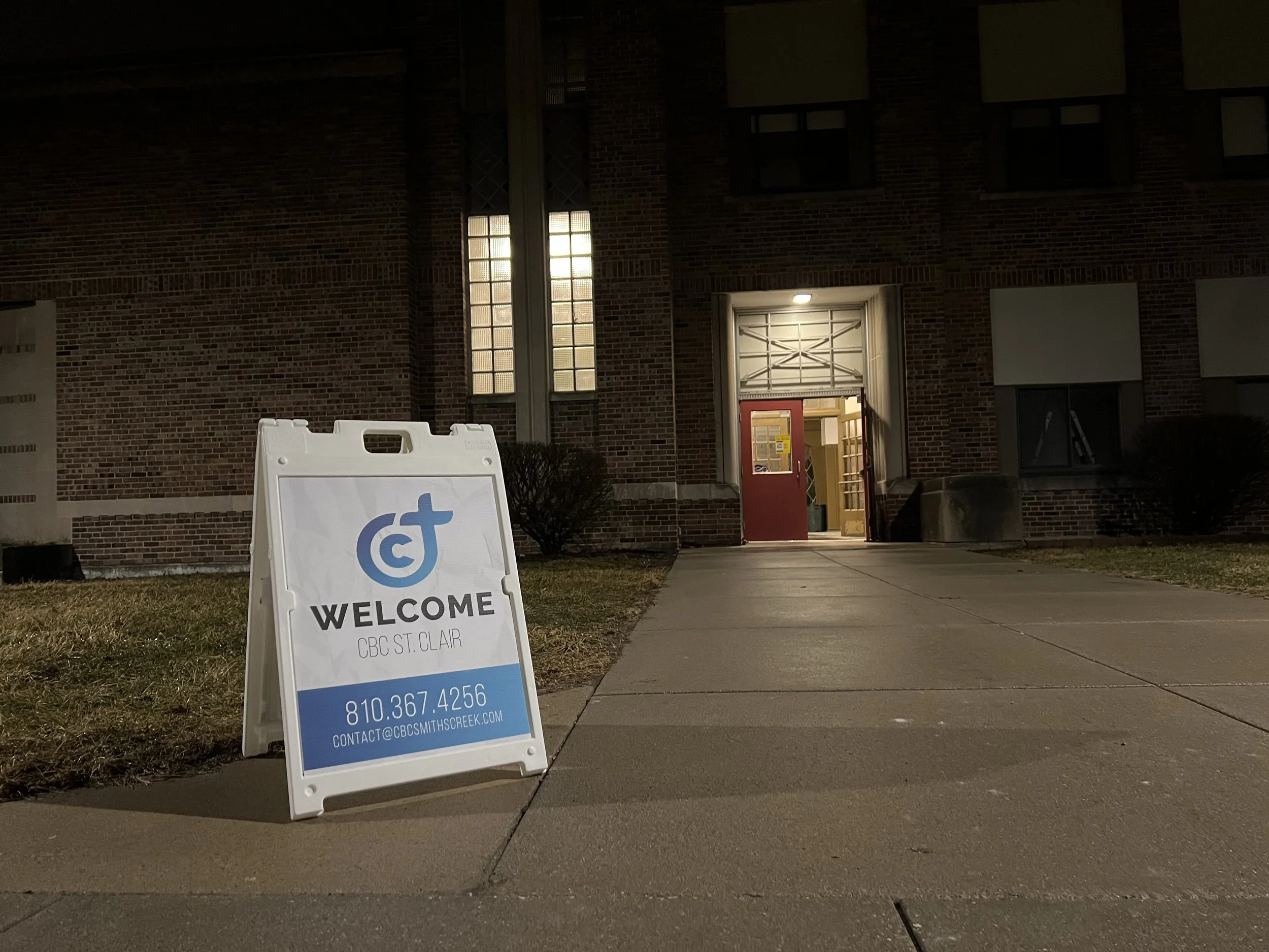 A sidewalk sign outside a brick building at night, welcoming visitors to CBC St. Clair with contact information and website, illuminated by nearby light.
