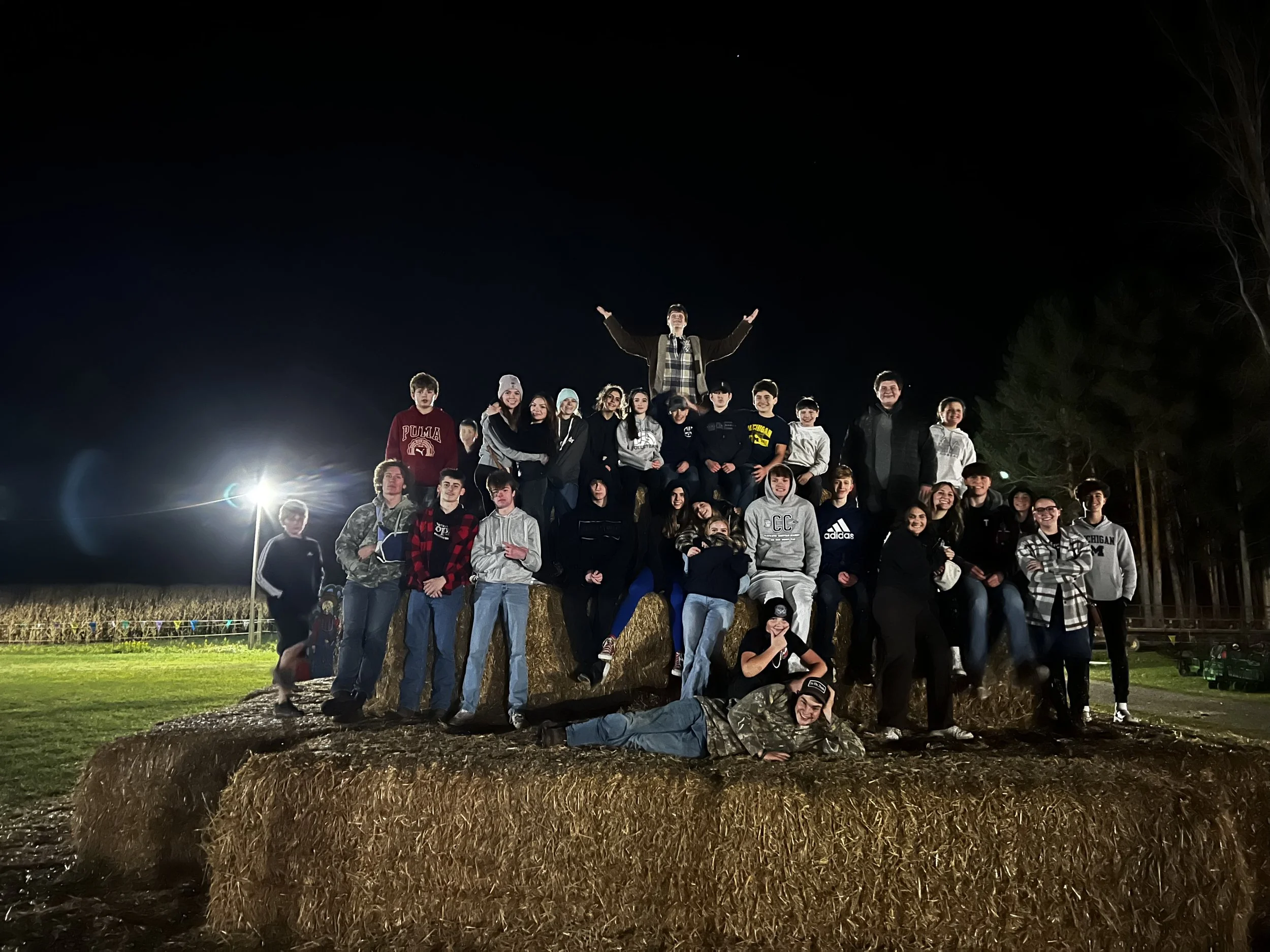 A large group of people, including children and teenagers, is gathered outdoors at night, standing on a hay bale with a straw mound in front. A man stands above them with his arms raised. There are trees in the background and bright lights illuminating the scene.